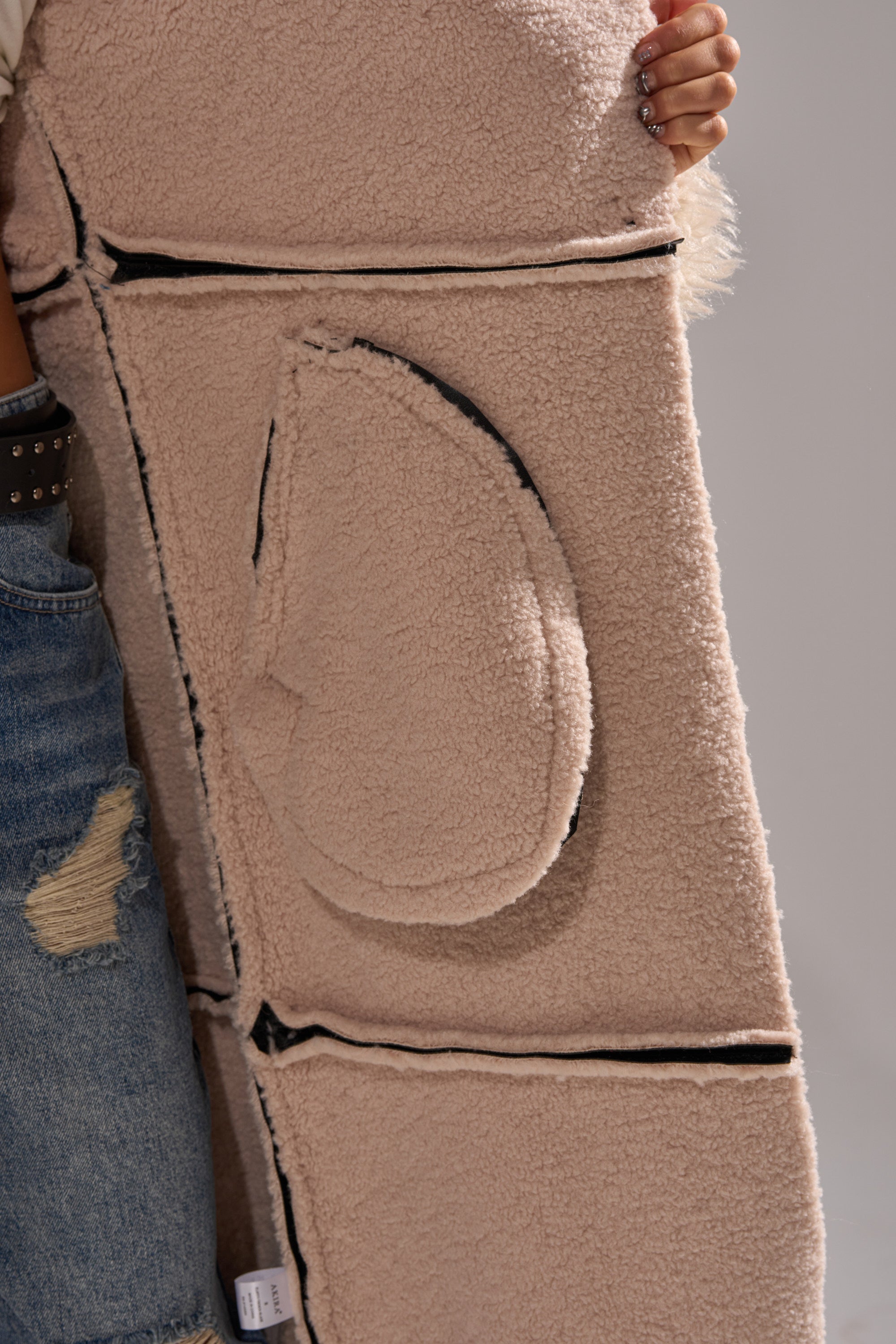 A person in ripped blue jeans holds the SHE GOT IT FLUFFY PU TRENCH WITH BONDED SHERPA, a beige, fleece-textured coat featuring black-edged seams and a large rounded pocket, with their hand resting on the top corner.