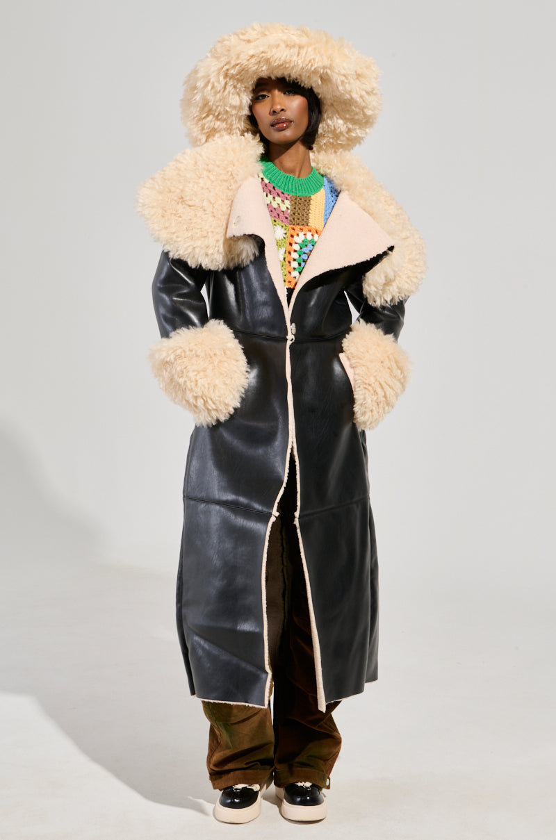 Dressed in the SHE GOT IT FLUFFY PU TRENCH WITH BONDED SHERPA, featuring a long black faux leather finish with a fluffy Sherpa collar and beige-trimmed cuffs, they accessorize with a large matching hat. Beneath the trench coat, they sport a colorful sweater with brown pants and complete the ensemble with black and white shoes. The background is plain gray.
