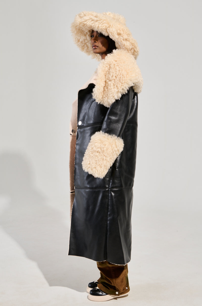 A person is wearing the SHE GOT IT FLUFFY PU TRENCH WITH BONDED SHERPA, featuring a long black leather-like finish with a fluffy sherpa collar and cuffs, accompanied by a wide-brimmed hat. Standing sideways, they showcase the faux sherpa lining that adds texture to the design. The plain light-colored background accentuates the trench coat's distinctive details.
