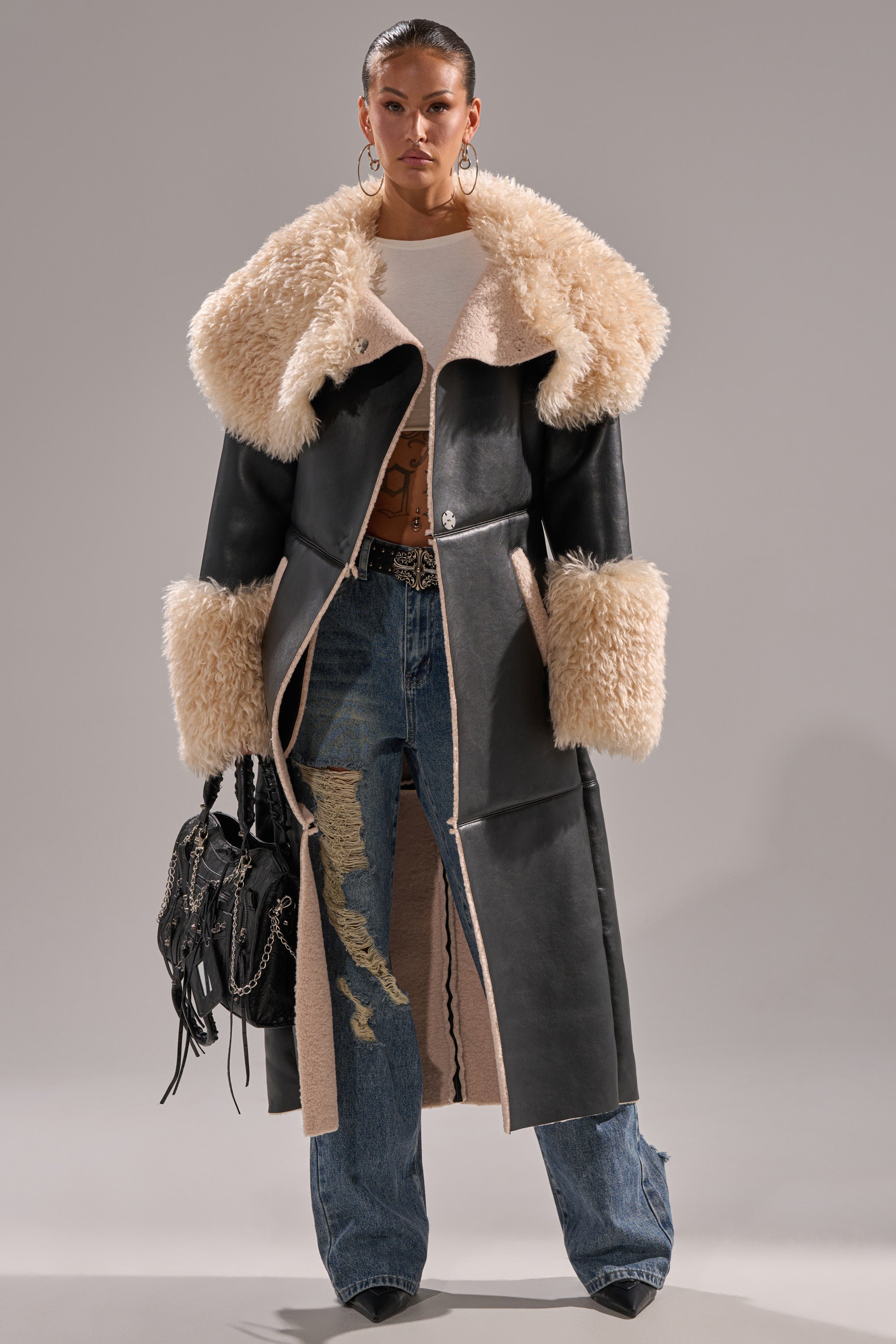 A woman faces forward in the SHE GOT IT FLUFFY PU TRENCH WITH BONDED SHERPA, featuring a large beige sherpa collar and cuffs, paired with distressed blue jeans, a beige top, hoop earrings, and a black fringed handbag.