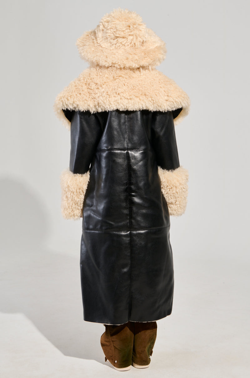 A person is wearing the SHE GOT IT FLUFFY PU TRENCH WITH BONDED SHERPA, featuring a long black design with a fluffy Sherpa collar, and beige fur accents on the cuffs and hood. Their back faces the camera against a plain background, complemented by brown shoes.