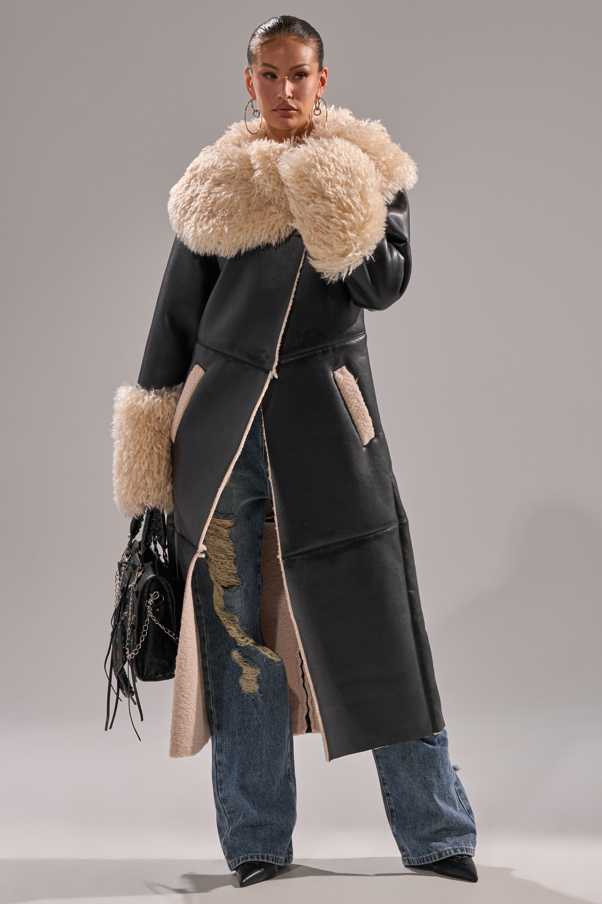 A person wearing the SHE GOT IT FLUFFY PU TRENCH WITH BONDED SHERPA, distressed blue jeans, pointed black shoes, hoop earrings, and holding a fringed black handbag stands against a plain gray background.