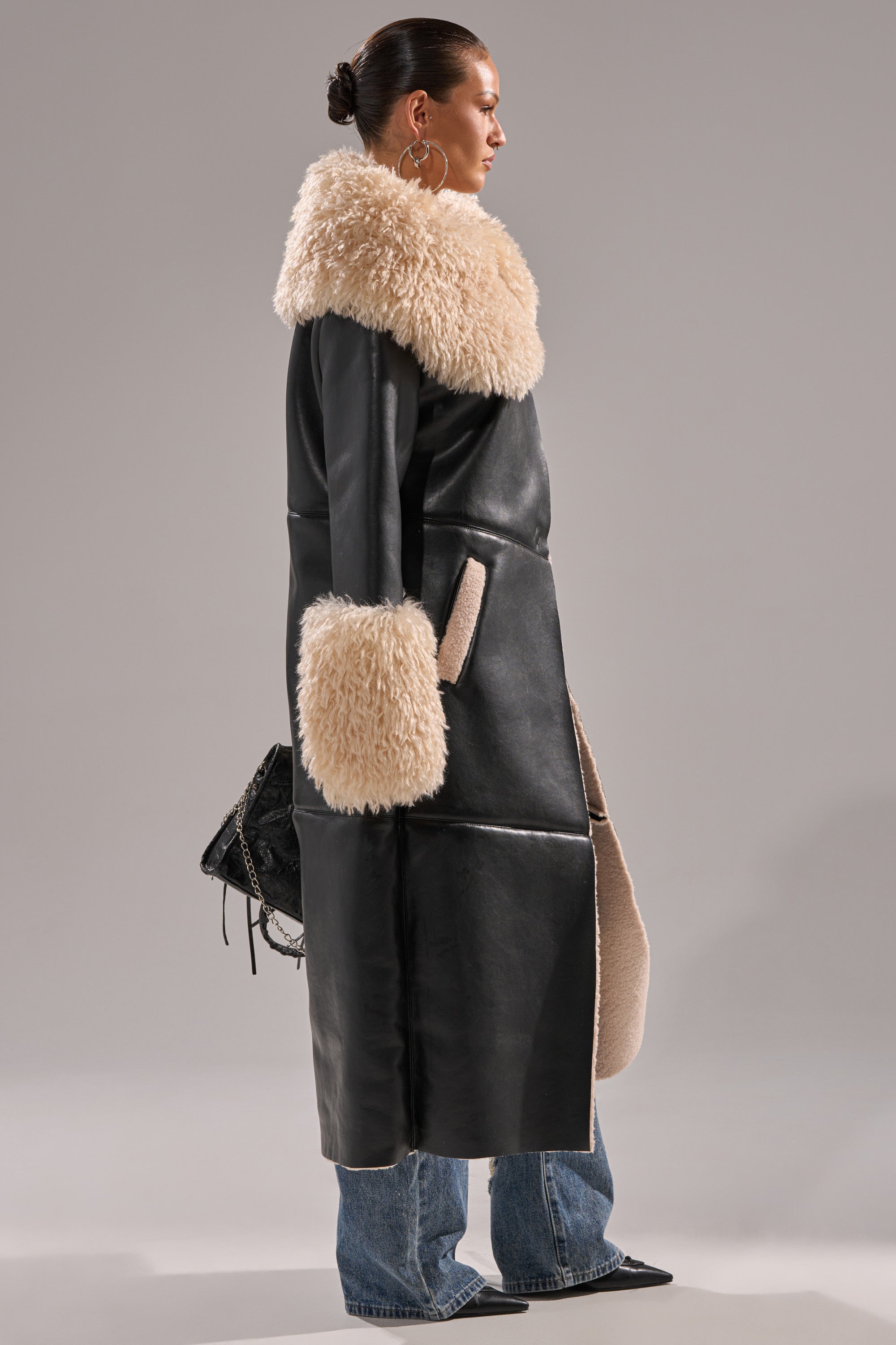 A woman stands in profile wearing the SHE GOT IT FLUFFY PU TRENCH WITH BONDED SHERPA—a long black coat with a large cream sherpa collar and cuffs—paired with blue jeans, black pointed shoes, and a black handbag.