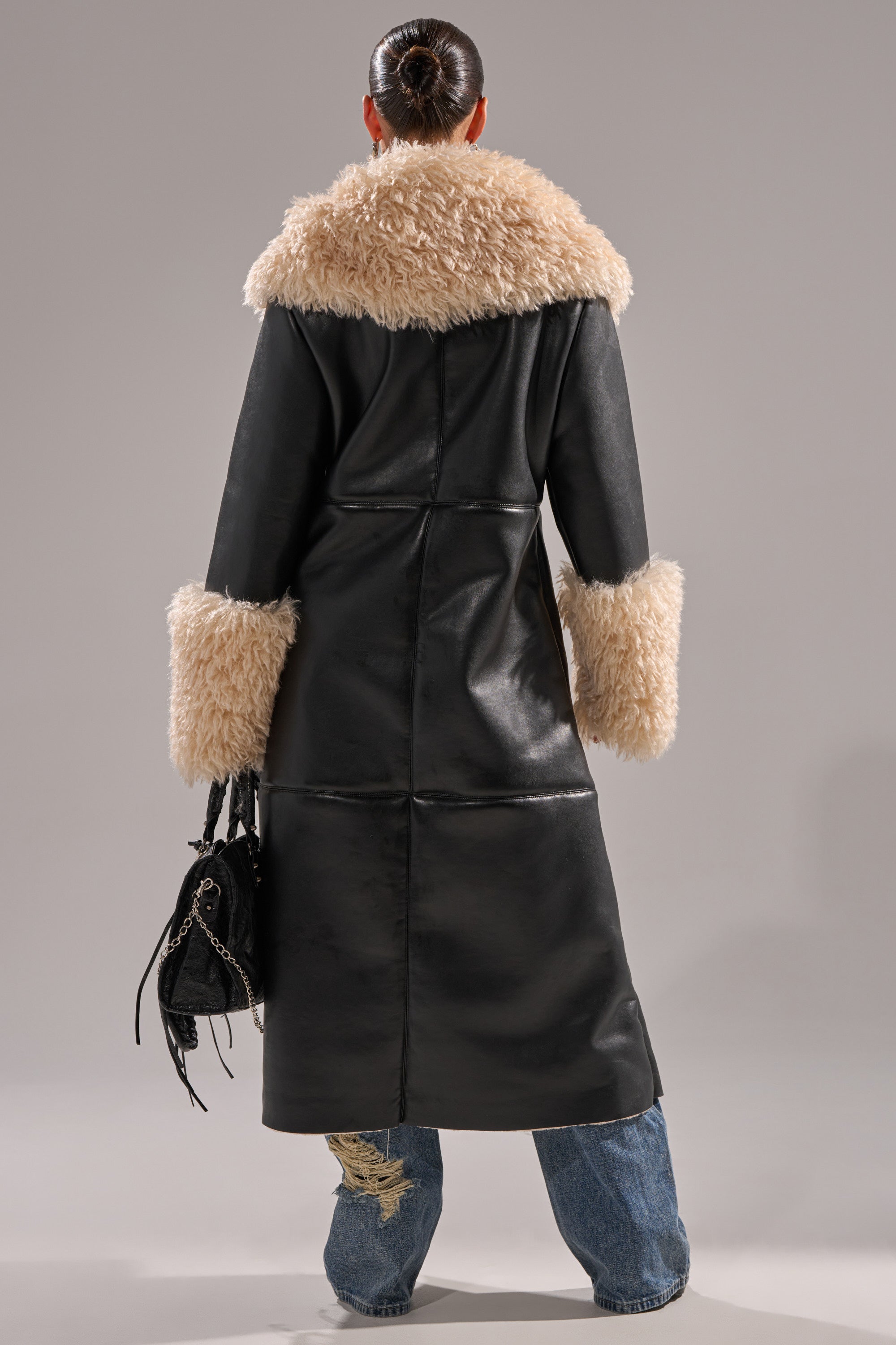 A person stands facing away, wearing the SHE GOT IT FLUFFY PU TRENCH WITH BONDED SHERPA, featuring faux leather and beige sherpa trim on the collar and cuffs, paired with blue jeans with a torn hem and a small black handbag.