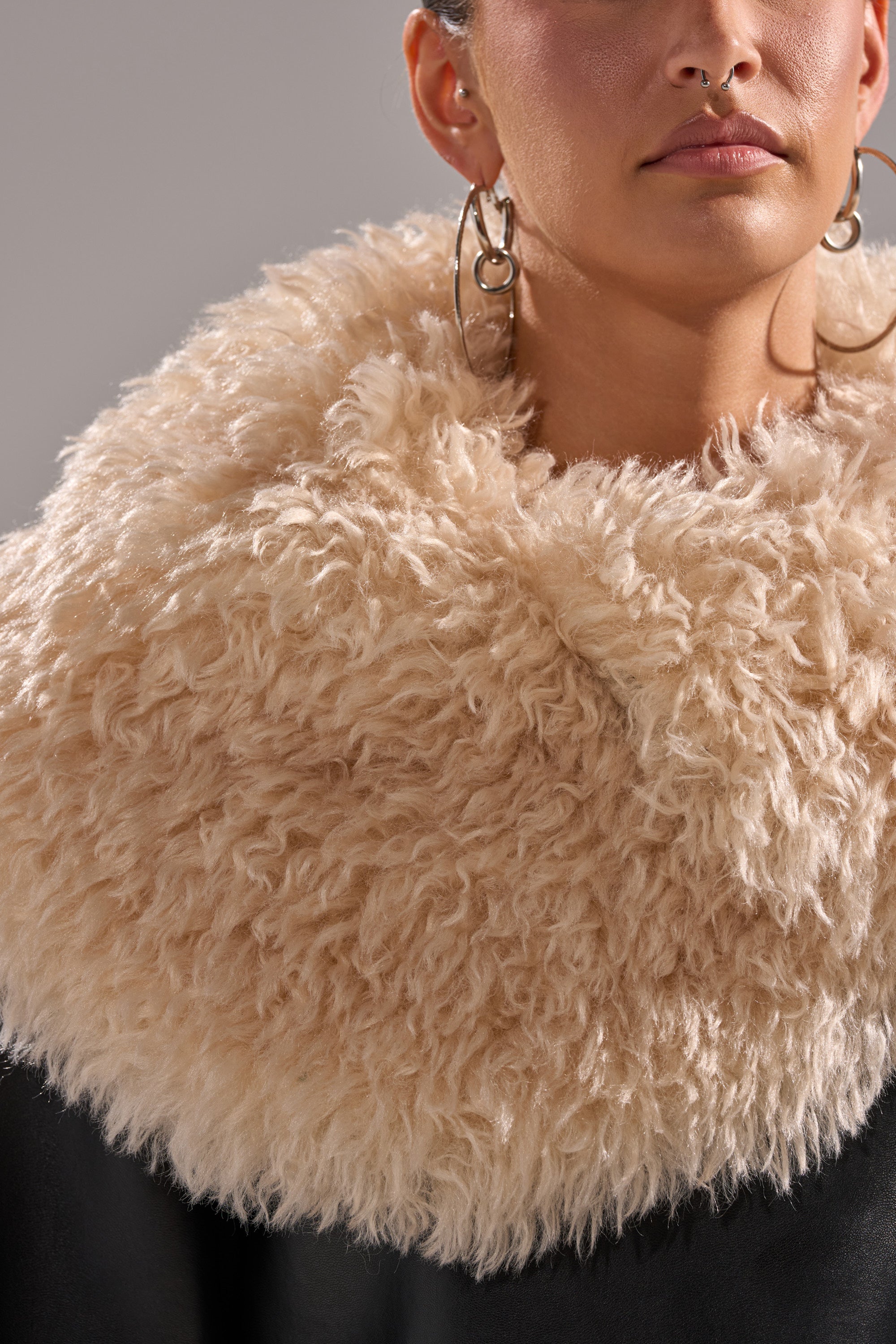 A woman wears the SHE GOT IT FLUFFY PU TRENCH WITH BONDED SHERPA over a dark AKIRA Label outfit, accessorized with large hoop earrings. Her thick, beige, fluffy collar stands out against the neutral gray background. Her face is partially visible.
