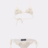 The CROCHET BEAUTY SWIMSUIT is a white boho-chic bikini set with floral appliqués and string ties at the neck, back, and hips, shown on a plain light background.
