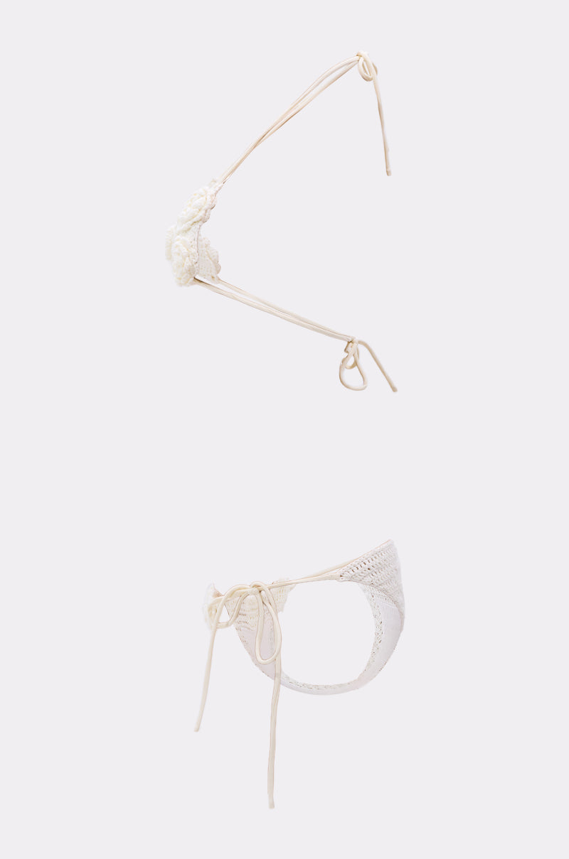 The CROCHET BEAUTY SWIMSUIT is a delicate white crochet set with a triangle top featuring floral appliqués and string ties, paired with matching bottoms with side tie closures, displayed on a plain light background.