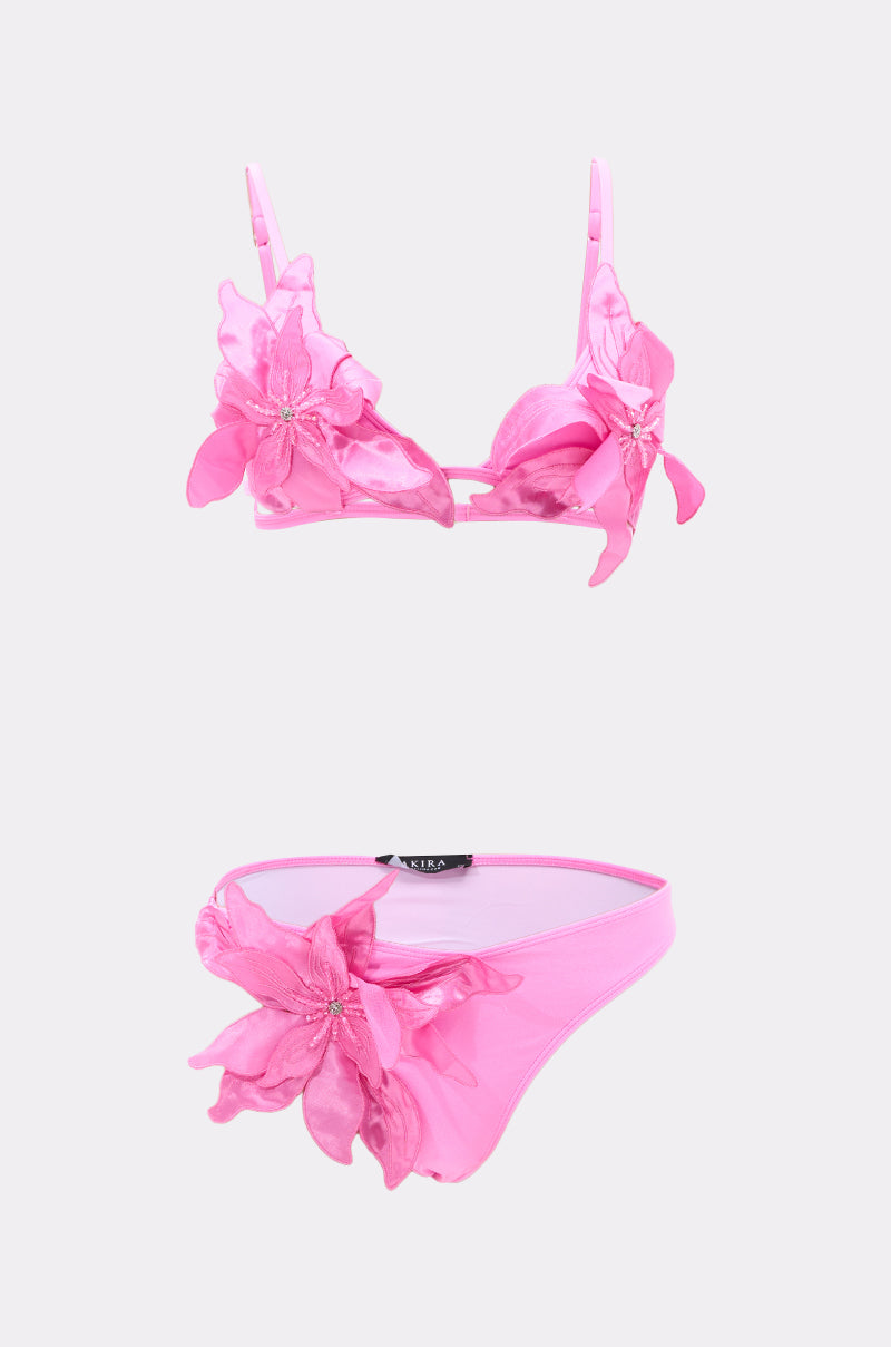 The IN BLOOM SWIMSUIT features a pink lingerie-inspired set with a bra and panties, both adorned with large sheer pink floral appliqués and delicate embellishments, shown on a plain light background.