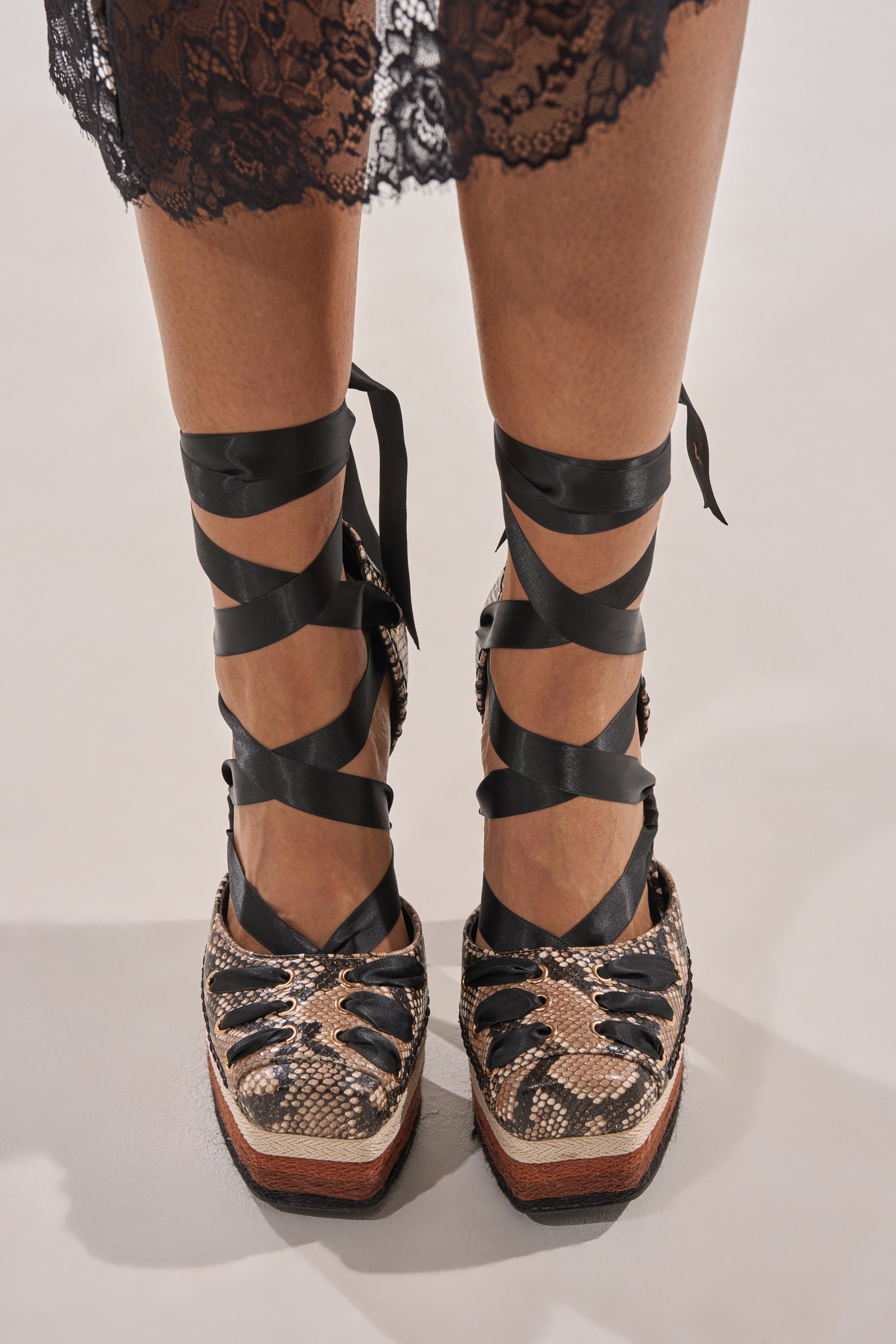 Wearing the AZALEA WANG FONTEYN SNAKE PLATFORM PUMP—black lace-up espadrille wedges with a snakeskin pattern and chunky block heel—paired with a black lace skirt. Photo taken from above, highlighting the shoes and lower legs. 3