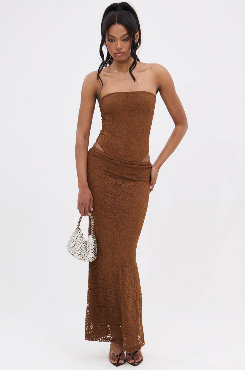 A woman stands against a white background wearing the PUCKER UP LACE MAXI SKIRT—a strapless, form-fitting brown dress with a sheer floral lace maxi skirt—paired with strappy heels and a small white beaded handbag.