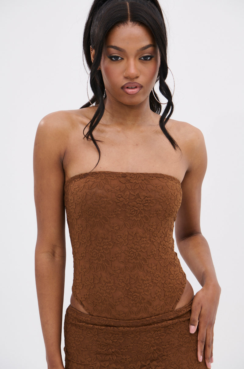 A woman with long dark hair in a high ponytail models the PUCKER UP LACE BODYSUIT, featuring a brown, strapless lace design. She poses against a plain white background with a neutral expression, looking slightly down.