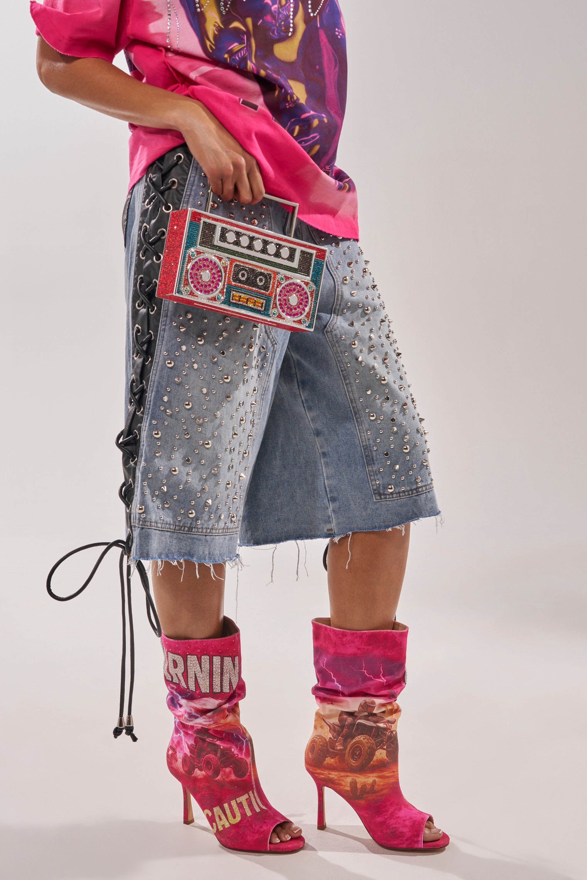 A person in a pink graphic tee, studded denim knee-length shorts with lace-up sides, a beaded cassette clutch, and the AKIRA GOLD FORMIDABLE PINK MULTI ABSTRACT OPEN TOE BOOTIE. 1