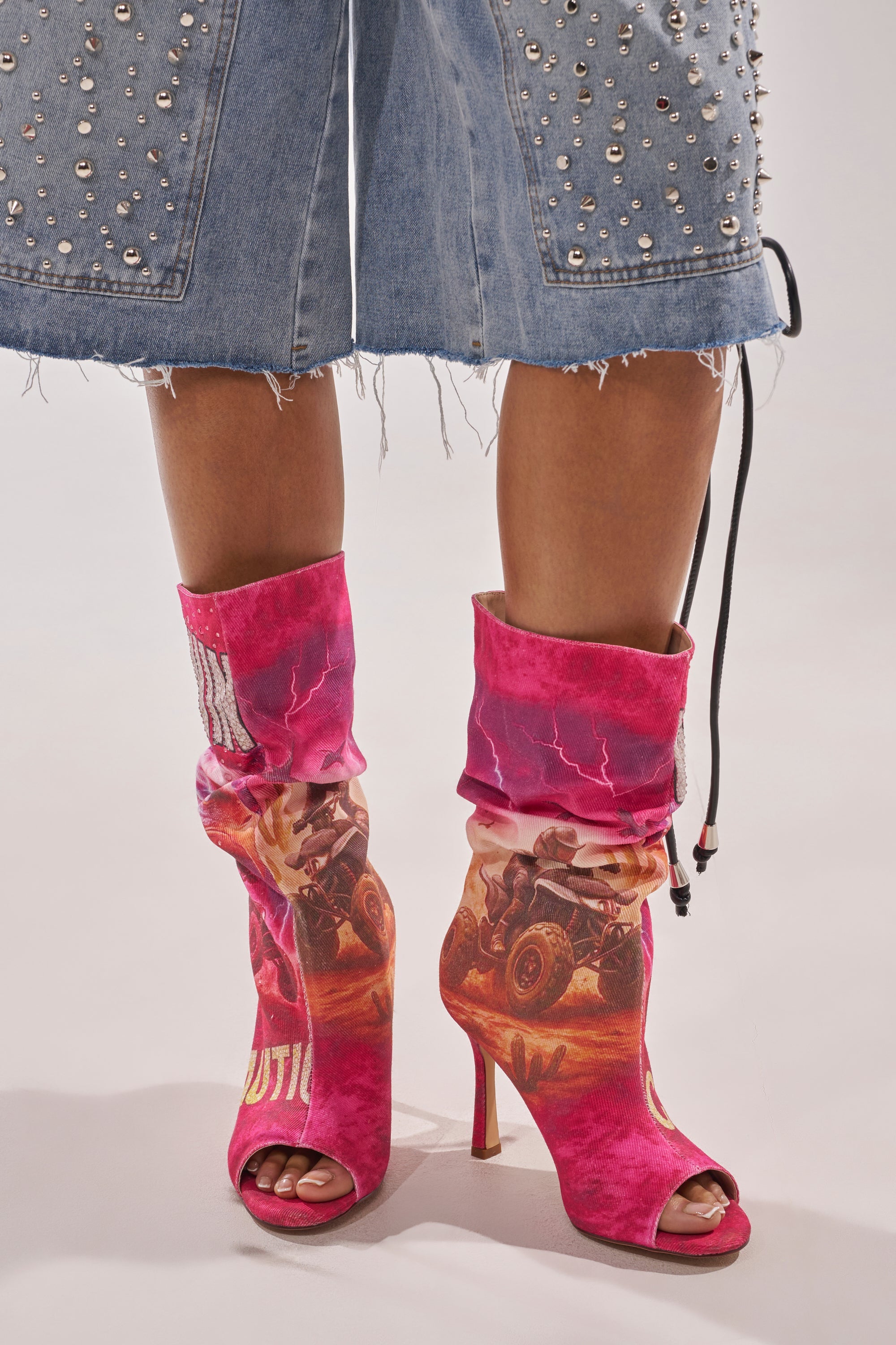 A person in knee-length, studded, frayed denim shorts and AKIRA GOLD FORMIDABLE PINK MULTI ABSTRACT OPEN TOE BOOTIE stilettos stands against a plain background. 2