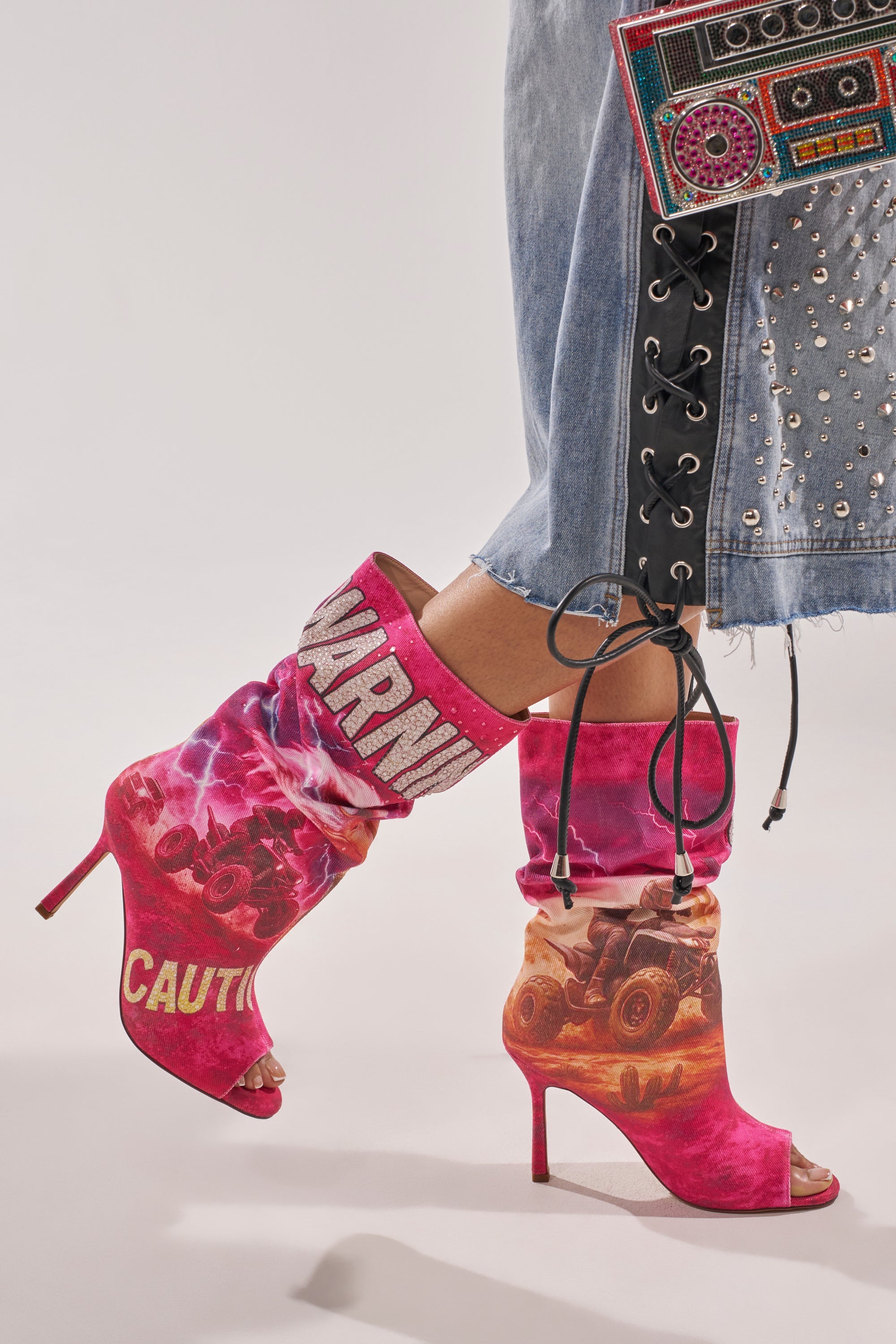 A person wears the AKIRA GOLD FORMIDABLE PINK MULTI ABSTRACT OPEN TOE BOOTIE, featuring pink high heels with “CAUTION,” “WARM,” and monster truck graphics. They accessorize with a colorful beaded clutch and a studded denim skirt. 0