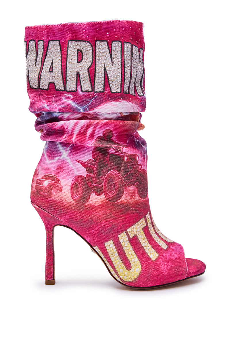 The AKIRA GOLD FORMIDABLE PINK MULTI ABSTRACT OPEN TOE BOOTIE features a mid-calf silhouette with slouchy pink fabric, bold “WARNING” and yellow “CAUTION” text, ATV rider and lightning graphics, plus a stiletto heel and open toe.