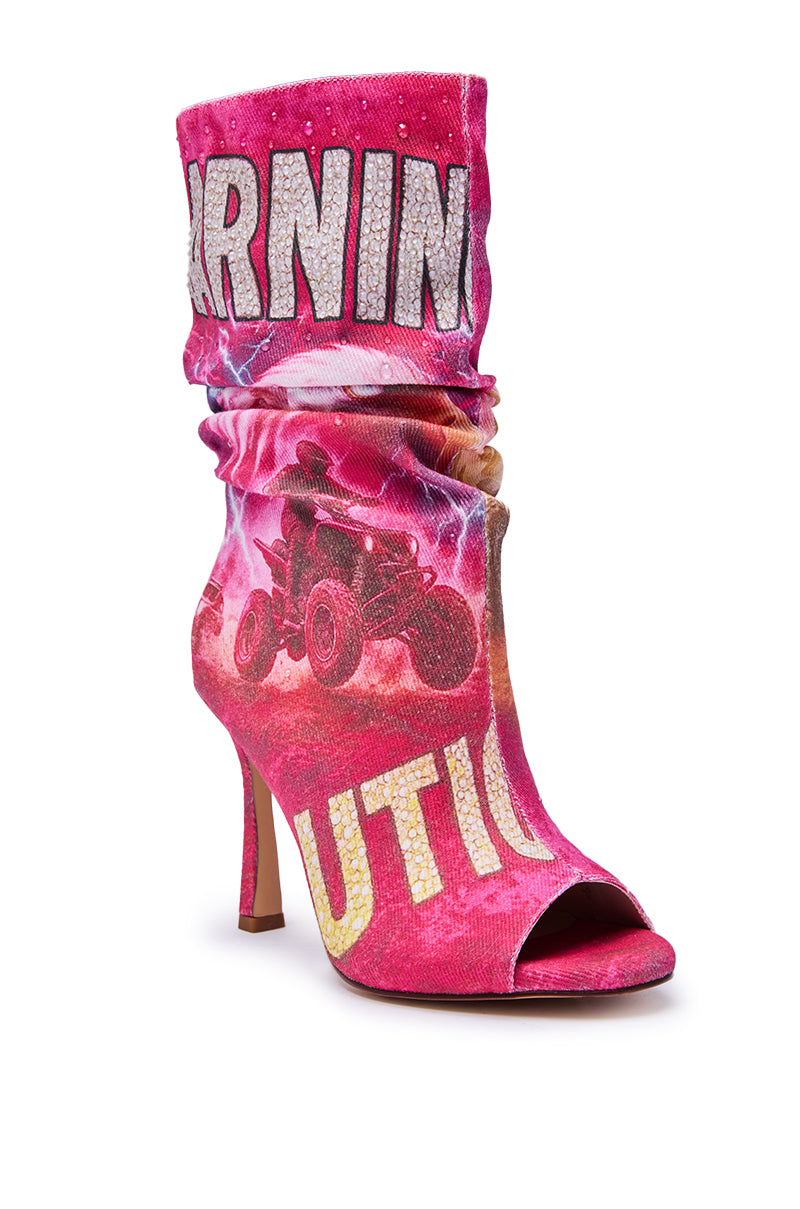 The AKIRA GOLD FORMIDABLE PINK MULTI ABSTRACT OPEN TOE BOOTIE is a high-heeled stiletto in pink, featuring bold graphics of a four-wheeler and large, partially visible "ARNING" and "UTION" text on a vibrant background.