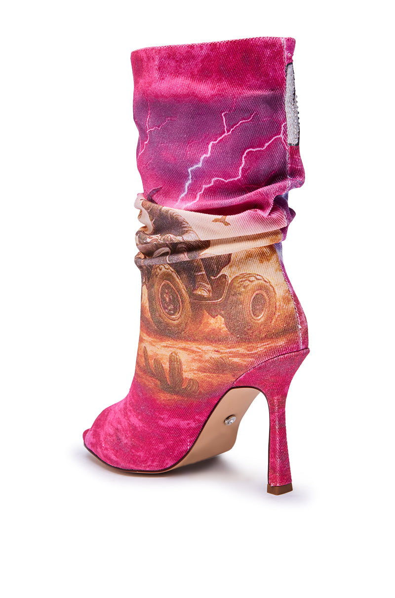 The AKIRA GOLD FORMIDABLE PINK MULTI ABSTRACT OPEN TOE BOOTIE features a high heel, vibrant pink fabric, and bold abstract designs with lightning bolts and a monster truck racing through a desert landscape.