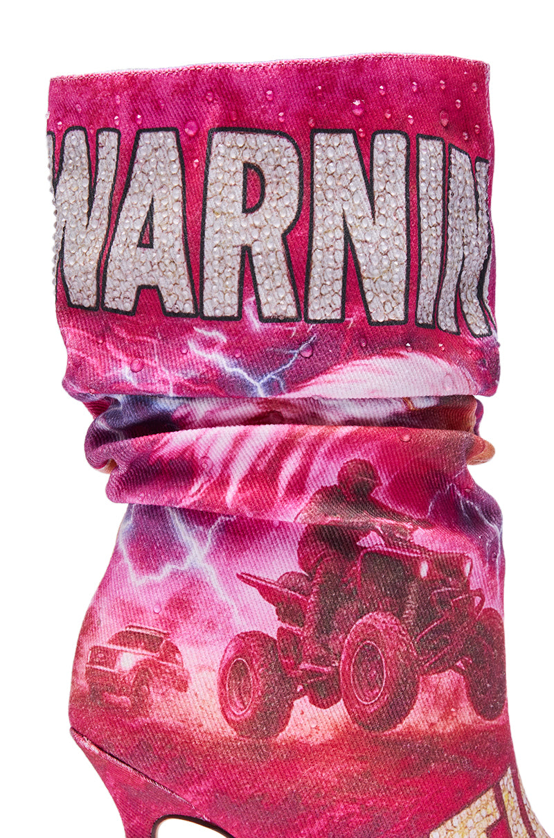 A close-up of the AKIRA GOLD FORMIDABLE PINK MULTI ABSTRACT OPEN TOE BOOTIE, featuring "WARNING" in bold sparkly letters and artwork of a person riding an ATV beneath a stormy, lightning-filled sky.