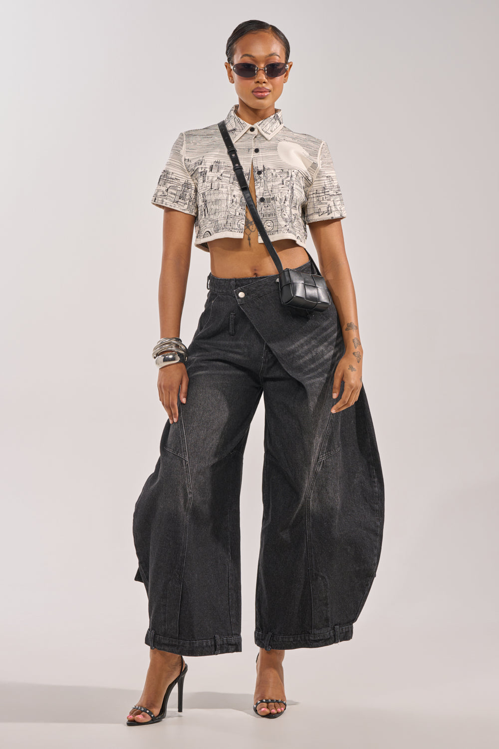 A woman in sunglasses poses confidently against a plain background, wearing oversized IZZY RELAXED DENIM IN BLACK WASH jeans with a cropped printed button-up shirt, black heels, a black crossbody bag, bracelets, and hoop earrings.