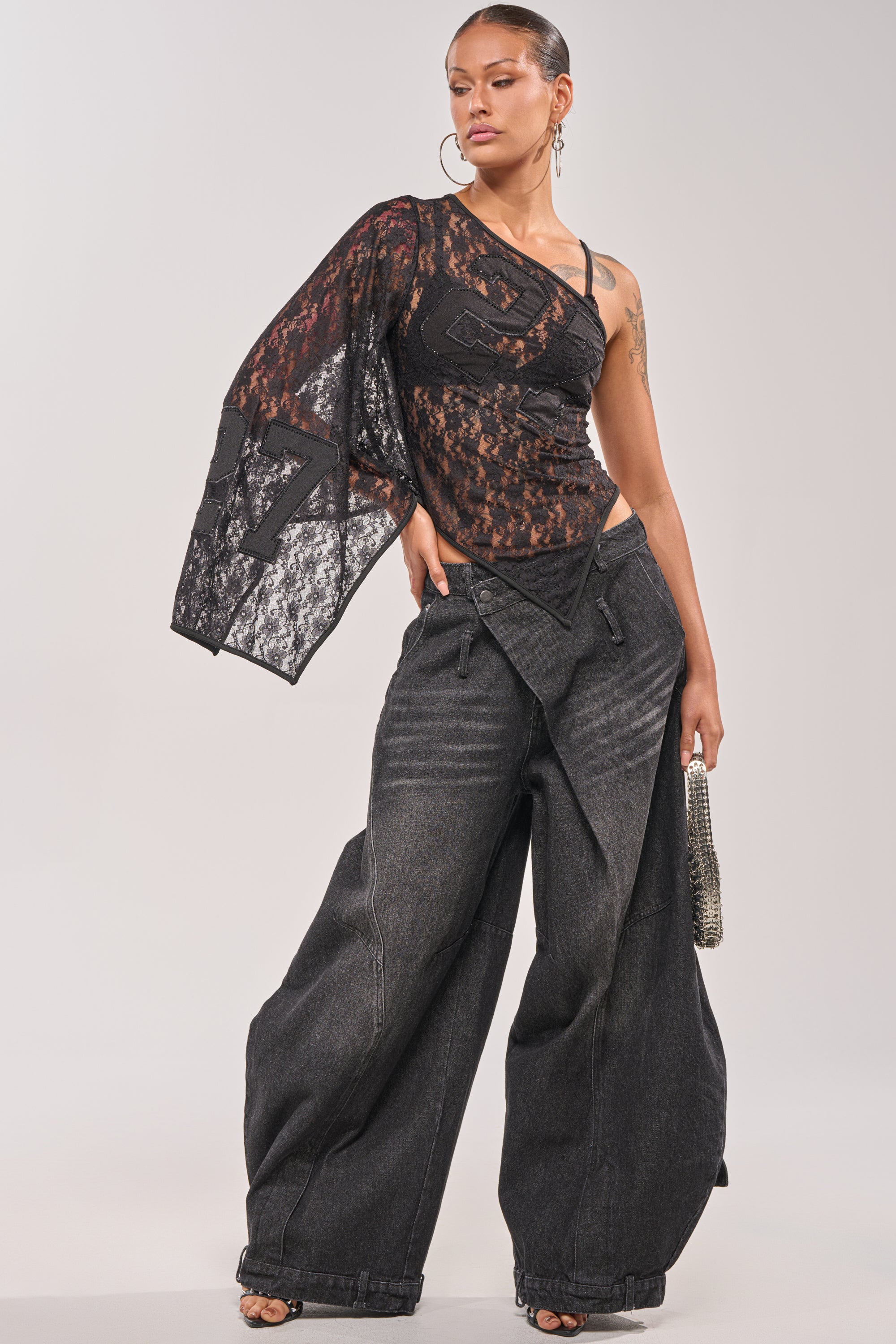 A woman poses confidently in IZZY RELAXED DENIM IN BLACK WASH wide-leg pants, styled with a sheer black asymmetrical lace one-sleeve top, small silver handbag, hoop earrings, and slicked-back hair. 0