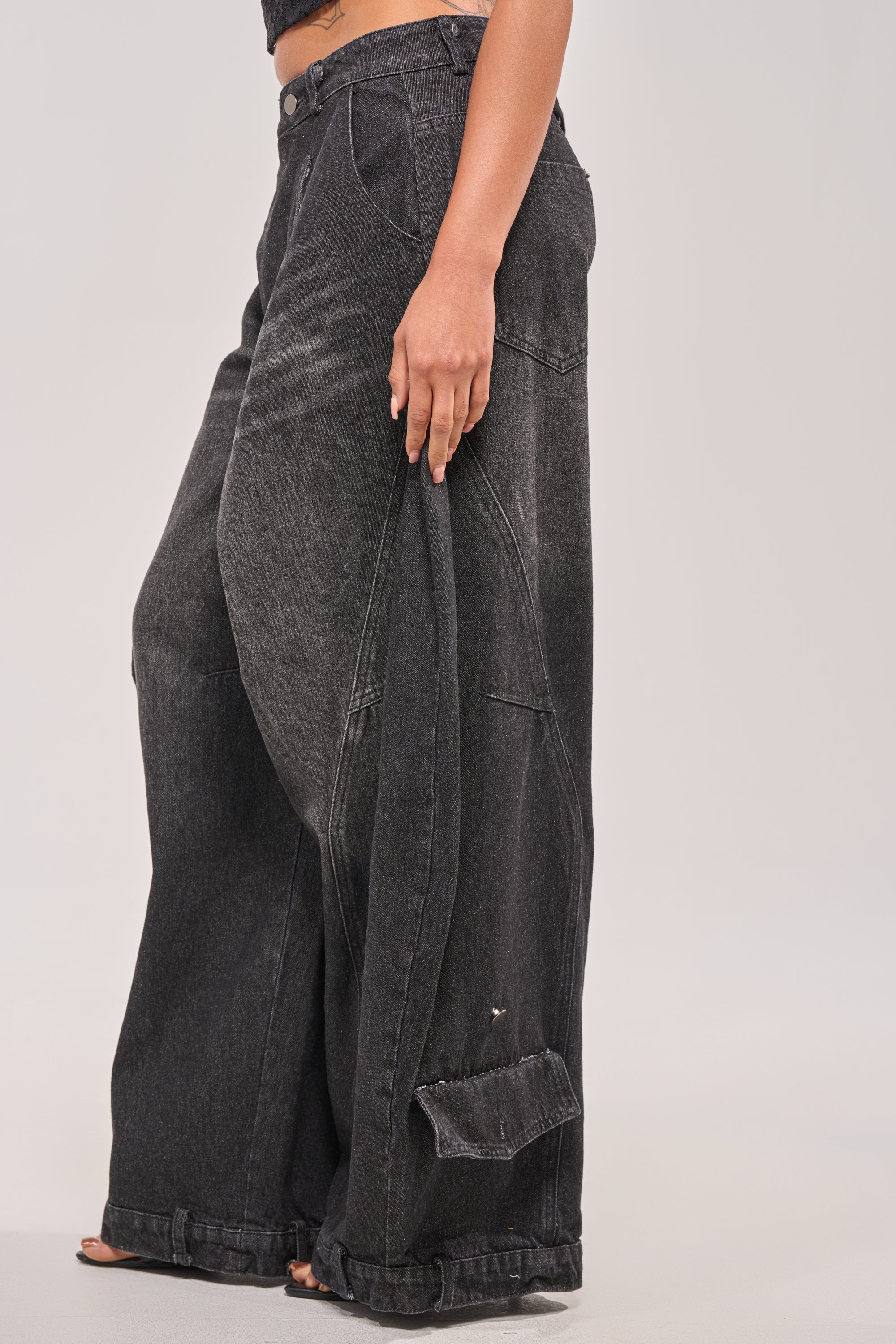 A person stands against a plain light background wearing the IZZY RELAXED DENIM IN BLACK WASH—ultra-baggy, high-rise jeans with visible seam details and an oversized fit that covers most of the feet. 3