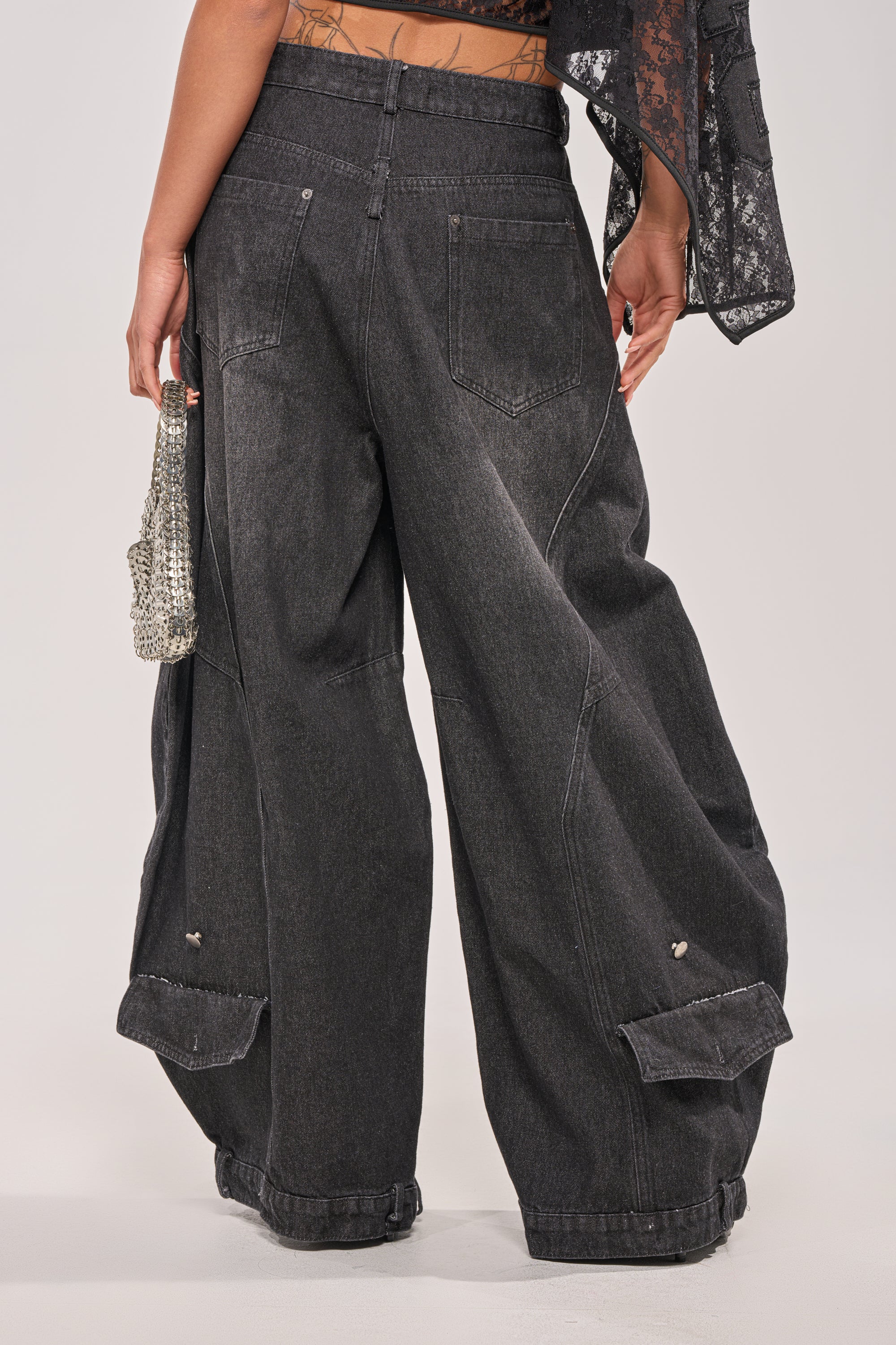 A person faces away, wearing the IZZY RELAXED DENIM IN BLACK WASH—ultra-baggy, high-rise black jeans with large back pockets and buttoned cuff flaps. They hold a silver purse, and part of a lacy black top is visible. 4