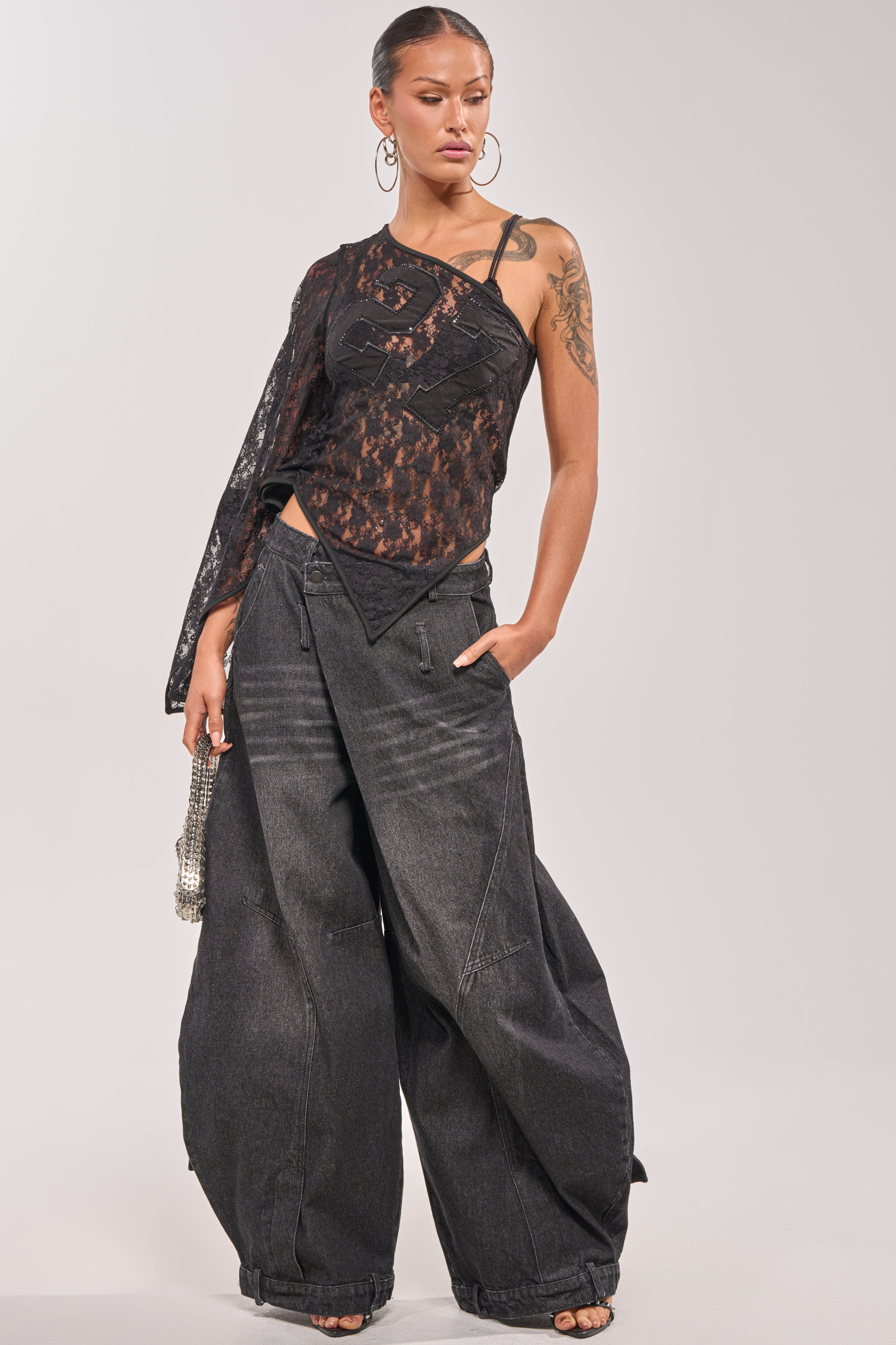 A woman poses in a black asymmetrical lace top and IZZY RELAXED DENIM IN BLACK WASH, accessorized with large hoop earrings and a silver handbag. Her hair is slicked back, and she casually rests one hand in her pocket. 5