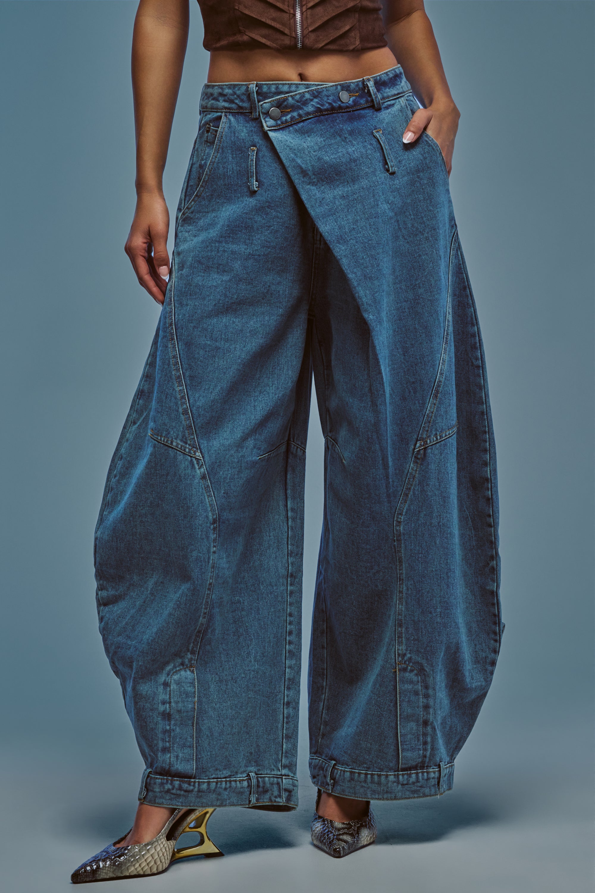 Wearing the IZZY RELAXED DENIM IN LIGHT BLUE WASH, a person showcases wide-leg barrel jeans with a unique folded front design. The look is styled with a brown top and pointed mesh-detailed shoes, set against a plain blue background.