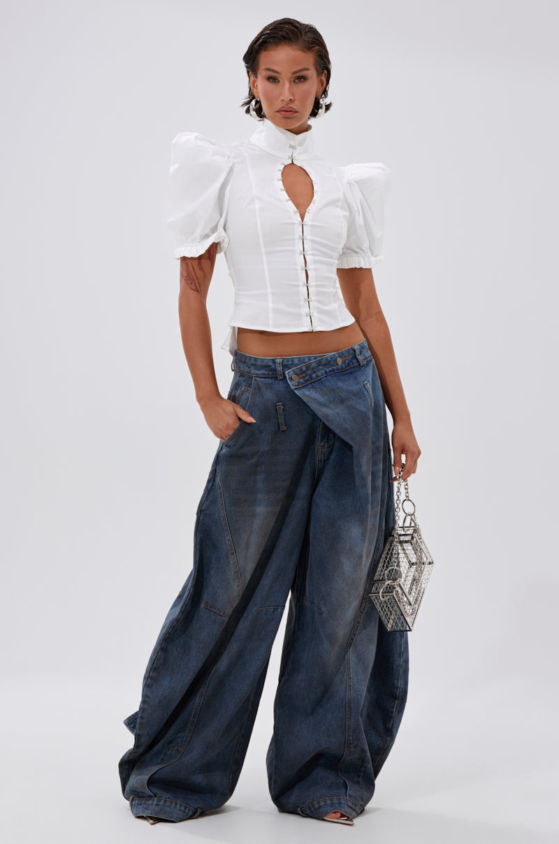 A woman wears a white puff-sleeve blouse with cut-out and buttons, paired with IZZY RELAXED DENIM high-rise jeans, holding a geometric silver handbag against a plain light background.