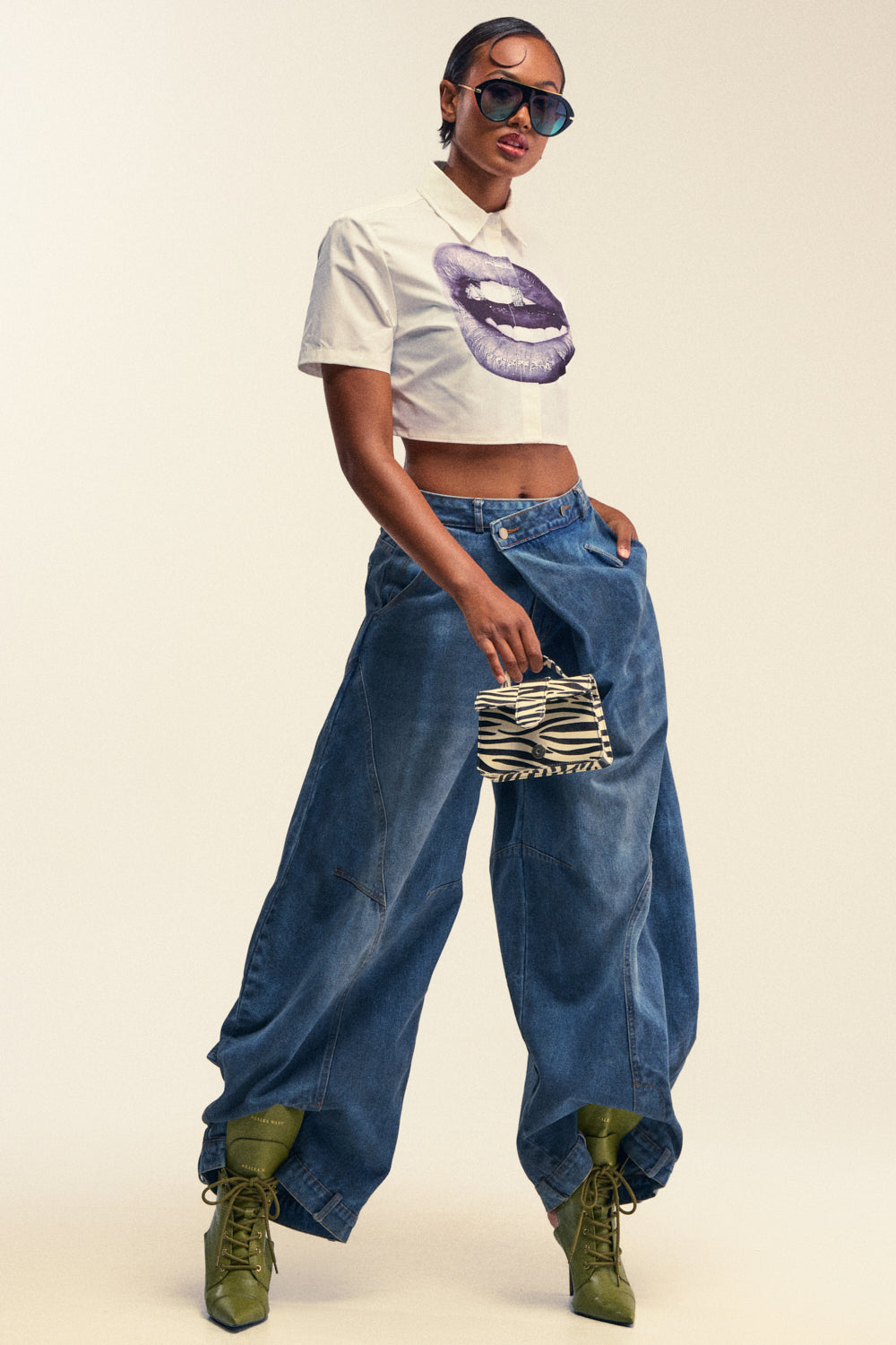 Wearing a white crop top with a purple lips graphic, the person pairs the look with IZZY RELAXED DENIM baggy wide leg jeans, green heeled boots, large sunglasses, and holds a small zebra print handbag while posing confidently against a plain background.