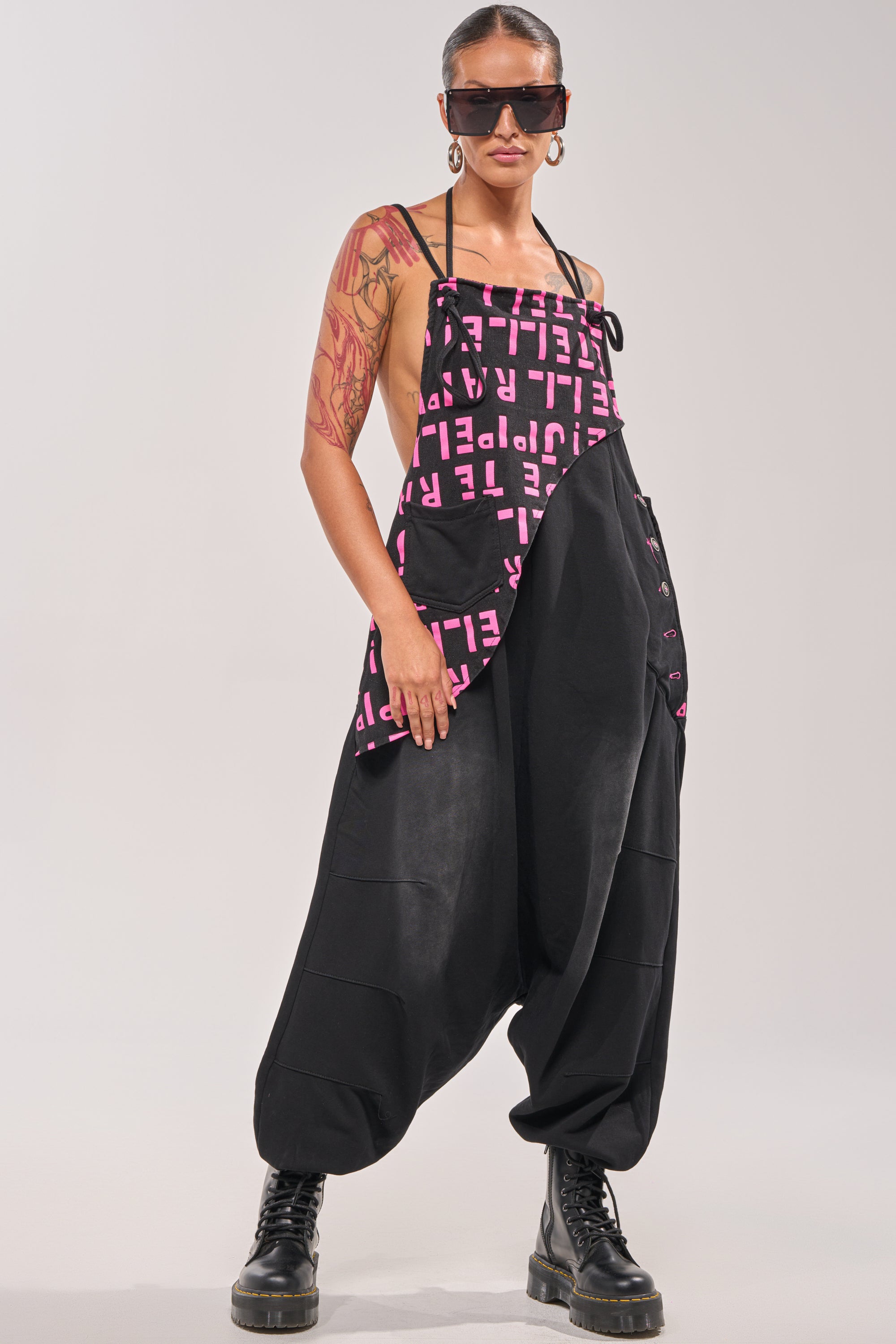 A person stands confidently against a plain backdrop wearing the WITH EASE GRAPHIC JUMPSUIT IN BLACK, chunky black boots, large hoop earrings, sunglasses, and showcasing tattoos on their right arm. 0