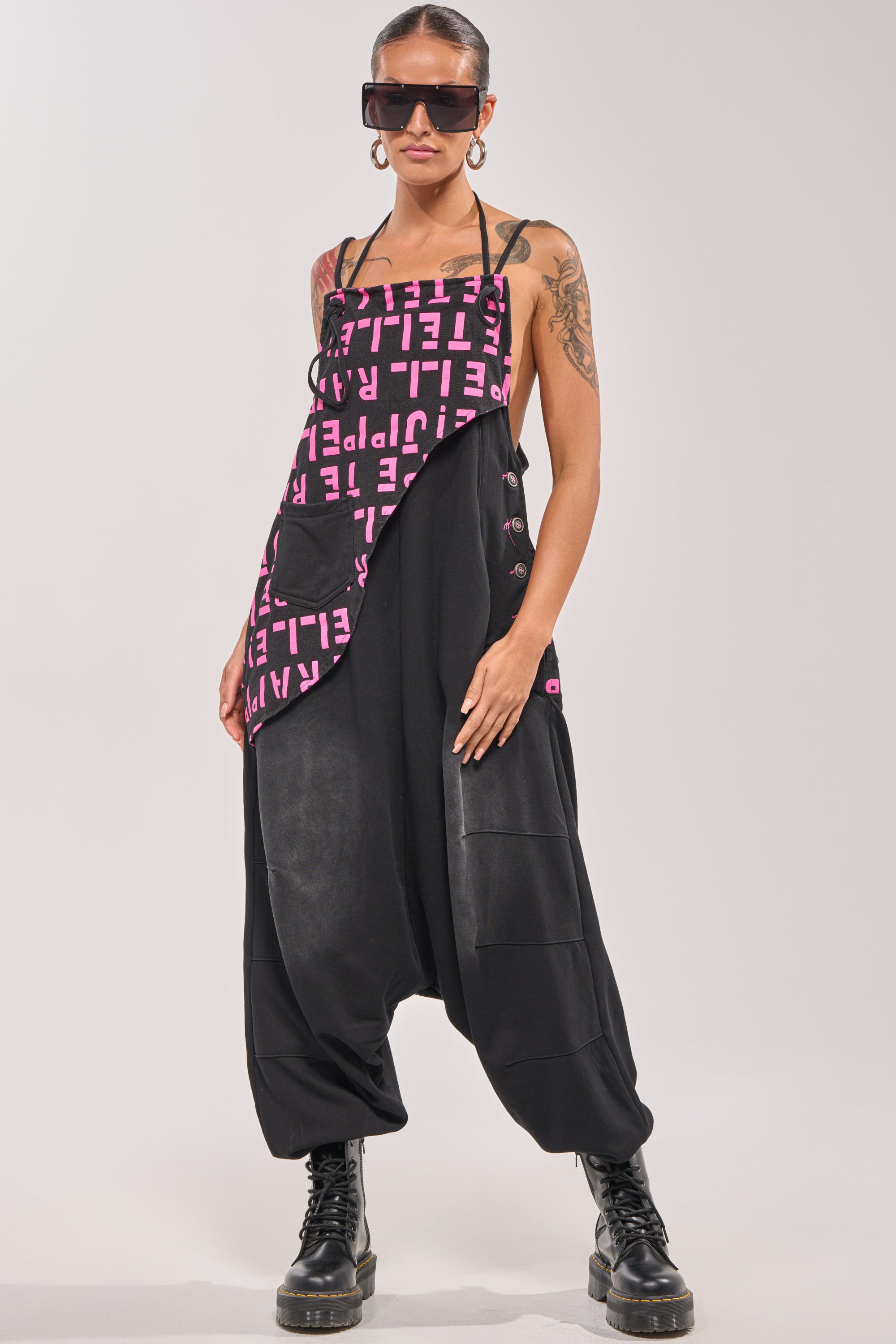 A person stands confidently against a plain background, wearing the WITH EASE GRAPHIC JUMPSUIT IN BLACK paired with oversized sunglasses and black combat boots. 1
