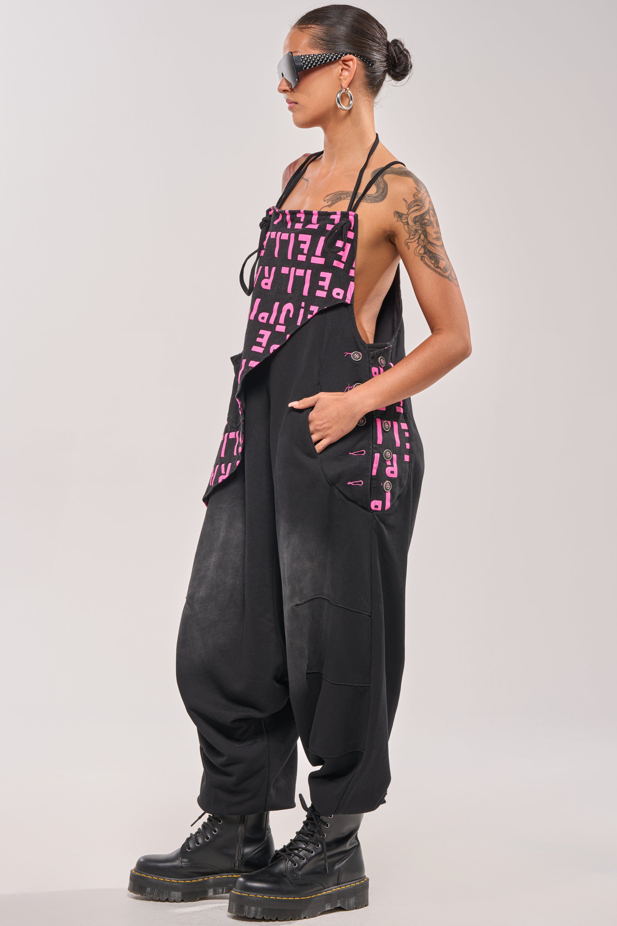 A person with a tattooed arm stands in profile against a plain light background wearing sunglasses, large hoop earrings, black boots, and the WITH EASE GRAPHIC JUMPSUIT IN BLACK. 4