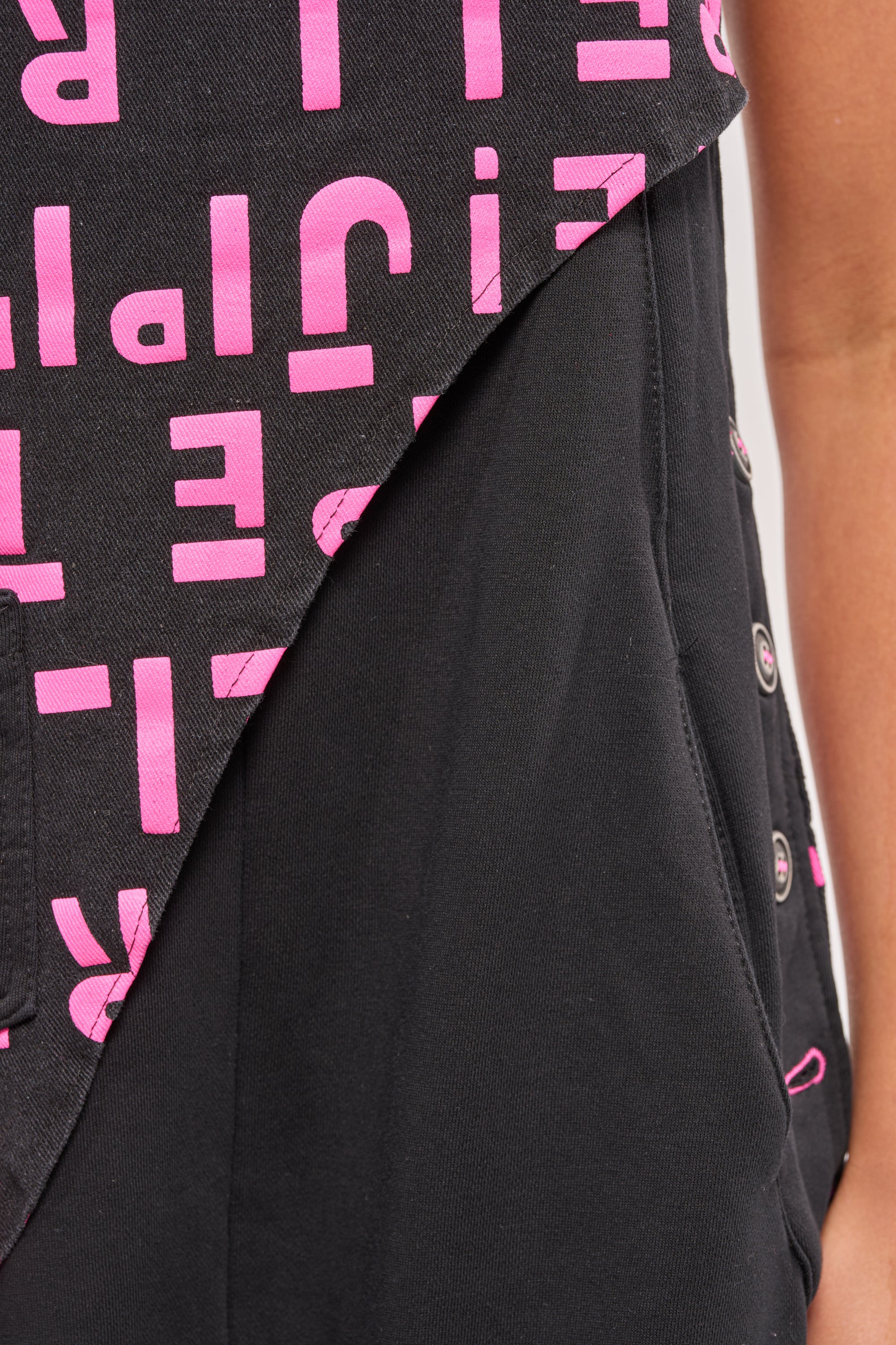 Close-up of a person wearing the WITH EASE GRAPHIC JUMPSUIT IN BLACK, featuring bold pink text print. The black jumpsuit has jogger-style details, with the person's arm visible on the right side of the image. 7
