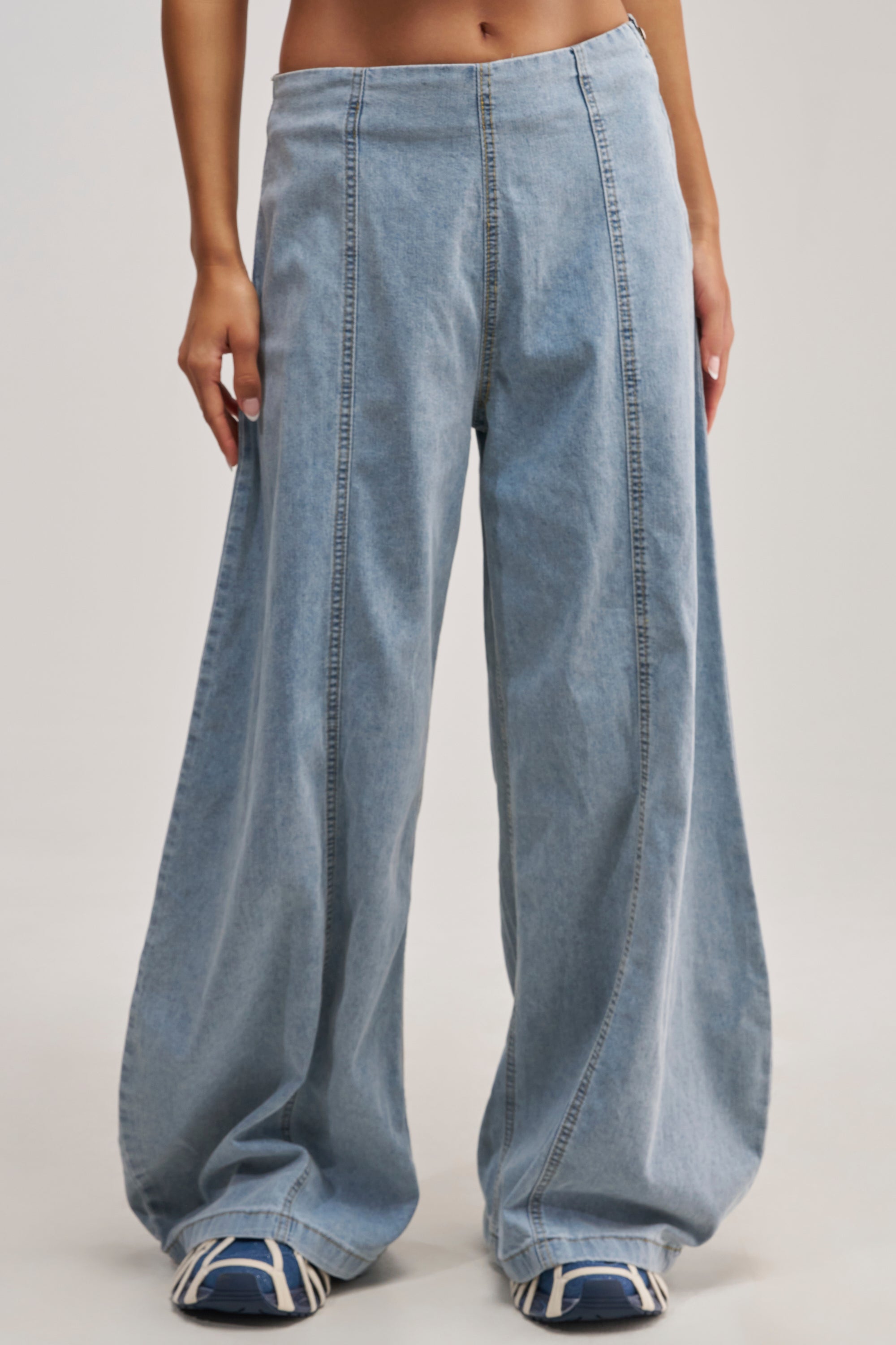 Person wearing the JUST IN TIME HIGH WAISTED RELAXED DENIM—loose, wide-leg light blue jeans styled with sneakers—standing against a plain light background. Only the lower torso, legs, and feet are visible.