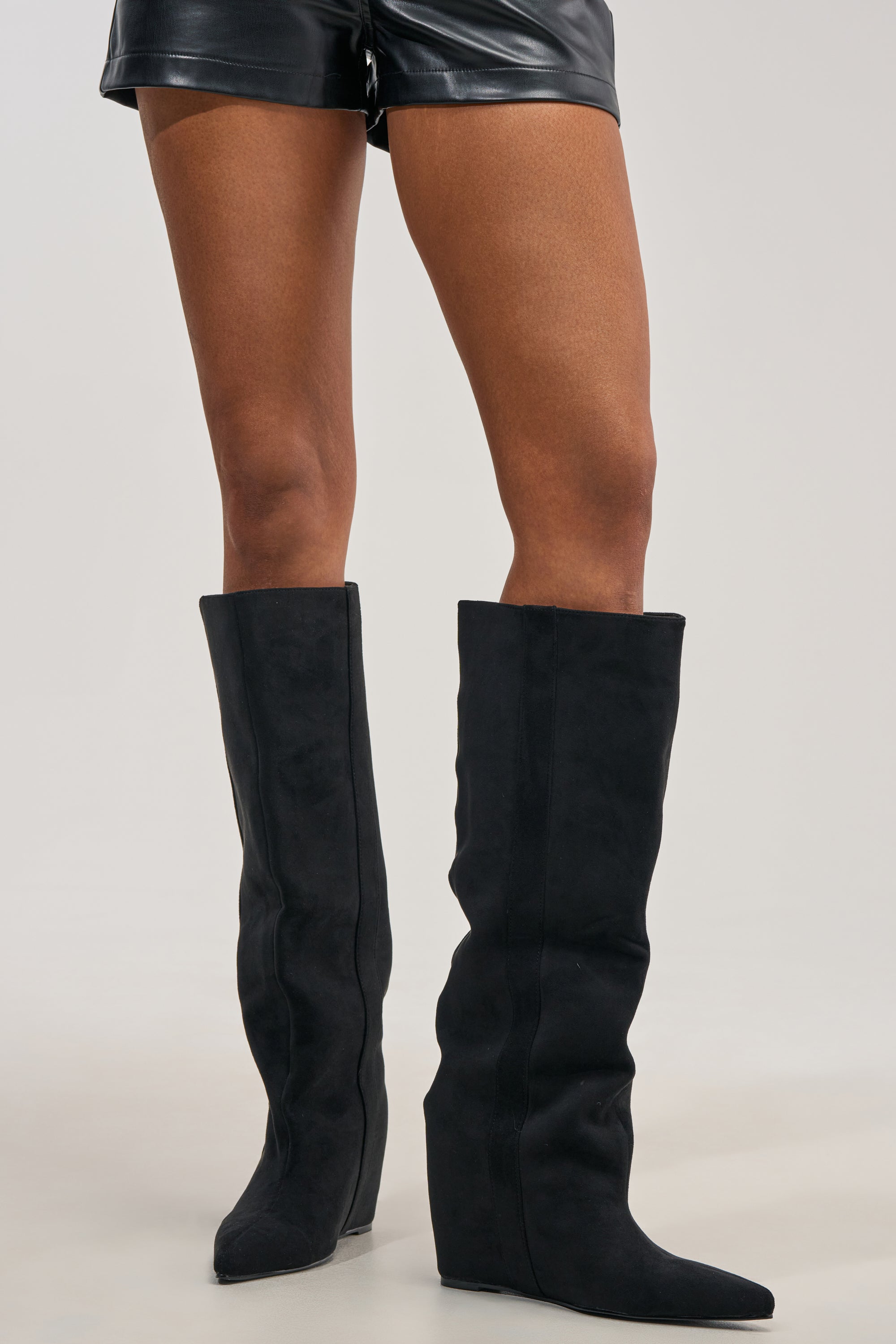 Wearing the AZALEA WANG FRANCES Black Faux Suede Hidden Wedge Boot paired with black leather shorts, a person stands against a plain light background.
