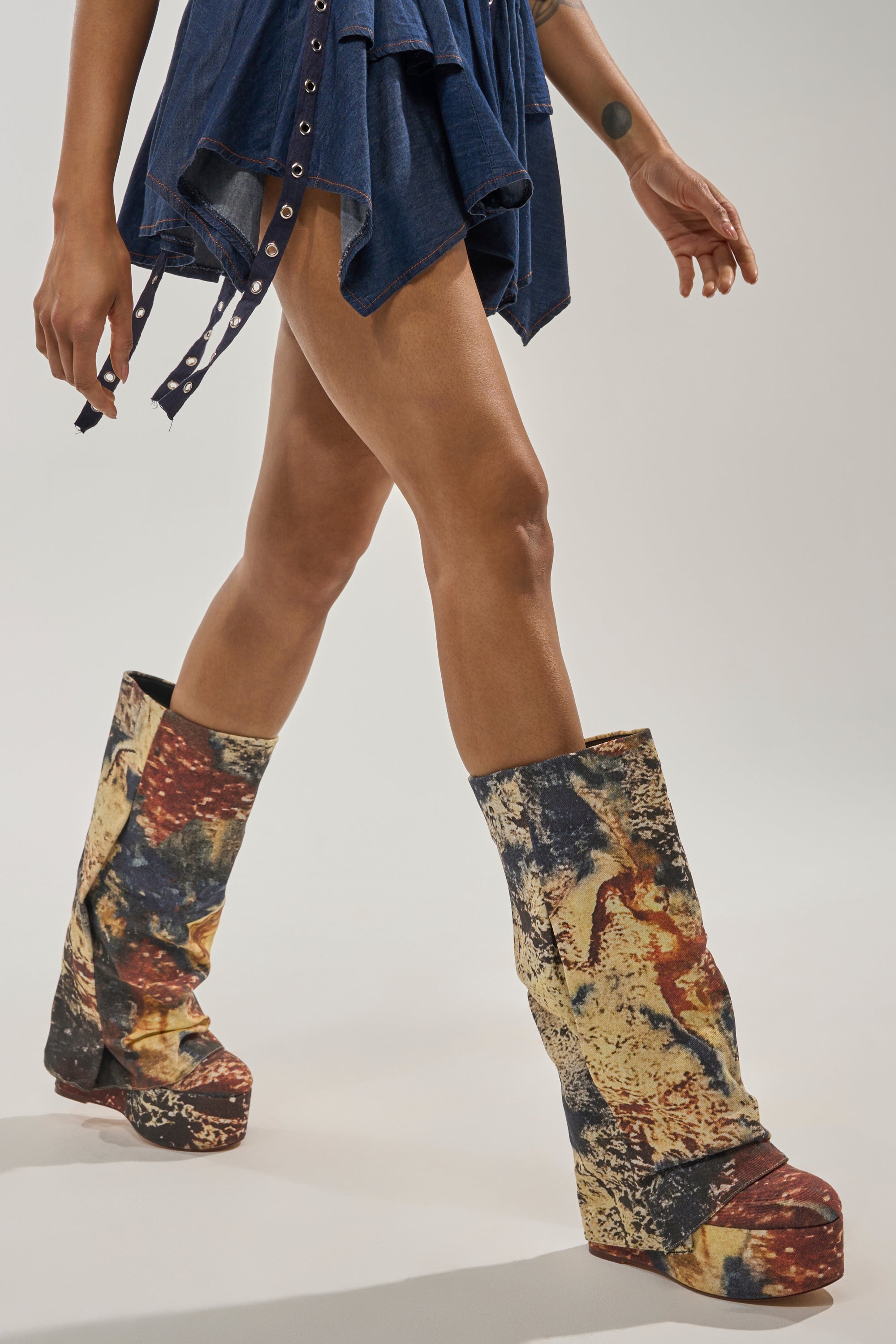 A person walks forward in a short blue ruffled dress, showcasing the AZALEA WANG FRANKEYE MULTI MATERIAL FLATFORM BOOT—knee-high with a multicolored abstract pattern. The image is cropped to reveal only their legs and part of the dress. 0