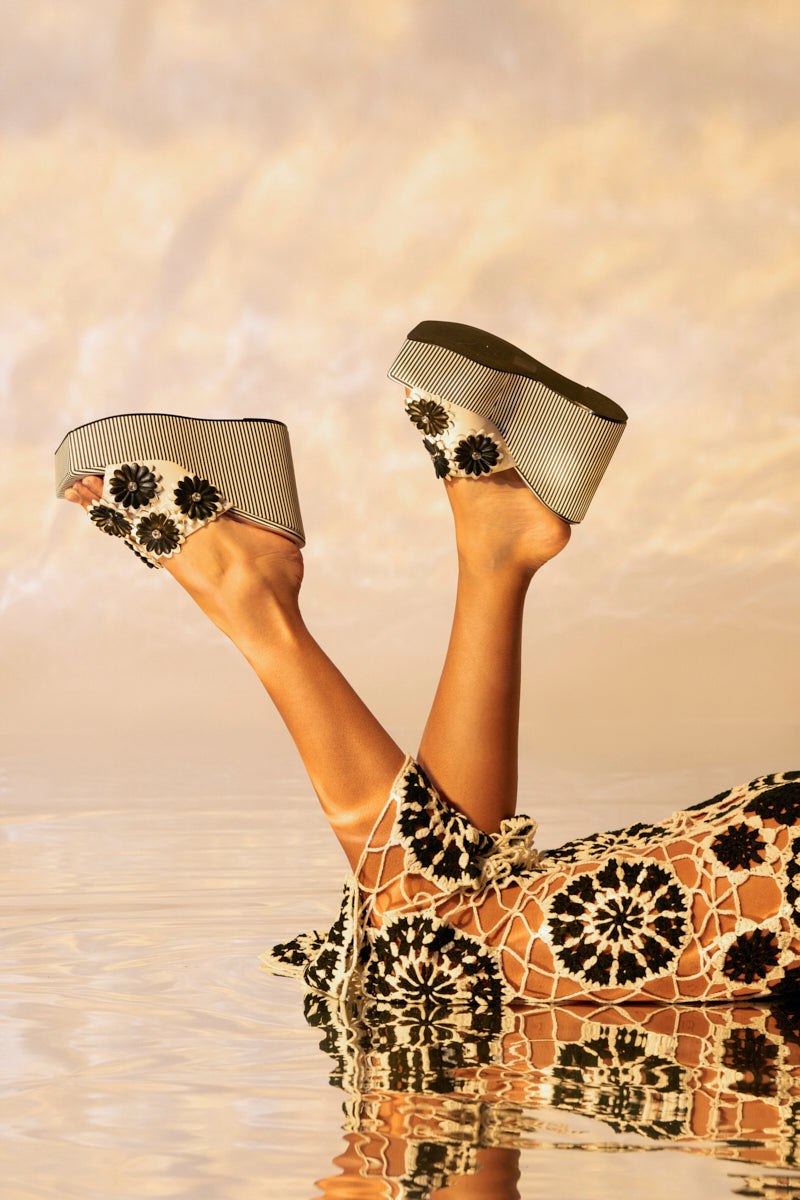A pair of legs rises above the water, highlighting AZALEA WANG FREA WHITE AND BLACK STRIPED FLORAL WEDGE SANDALS with cushioned insoles, complemented by black and white crocheted pants featuring a floral design, set against a warm, blurred background.