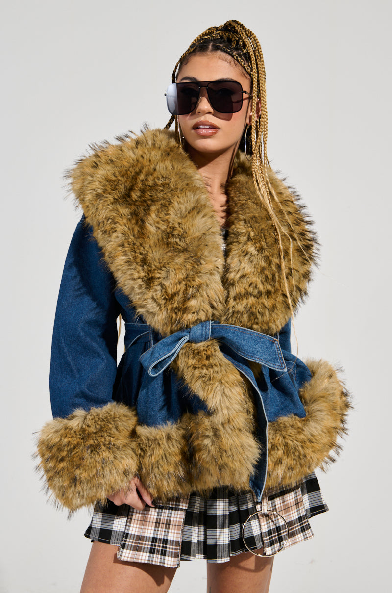 A person dons a FREDDIE FUR TRIM DENIM MOTO with its large faux fur collar and cuffs, paired with a plaid skirt. They sport sunglasses and long braids against a plain background, completing the look with an adjustable belt closure for added style.