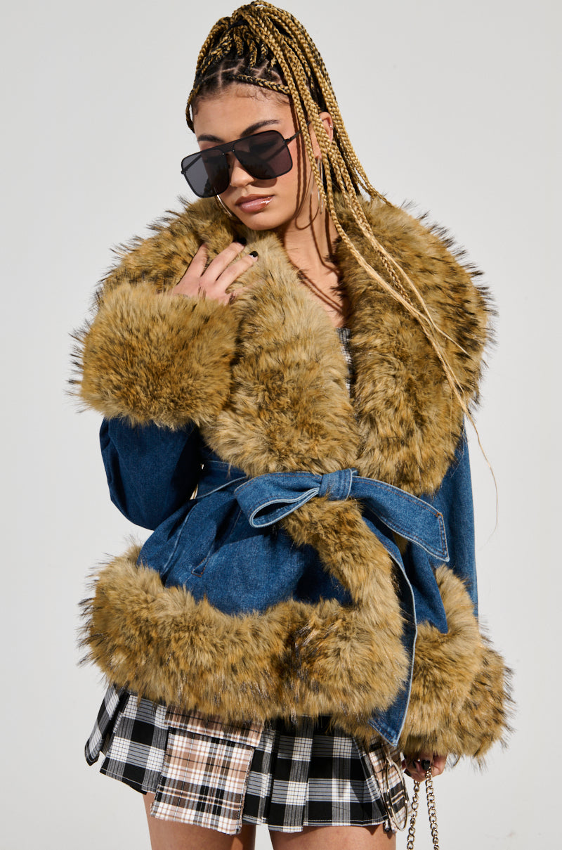 A person with braided hair wears oversized sunglasses and a FREDDIE FUR TRIM DENIM MOTO jacket featuring a brown faux fur trim. Paired with a plaid skirt, they strike a pose, hand confidently resting on the fur collar against a plain backdrop.