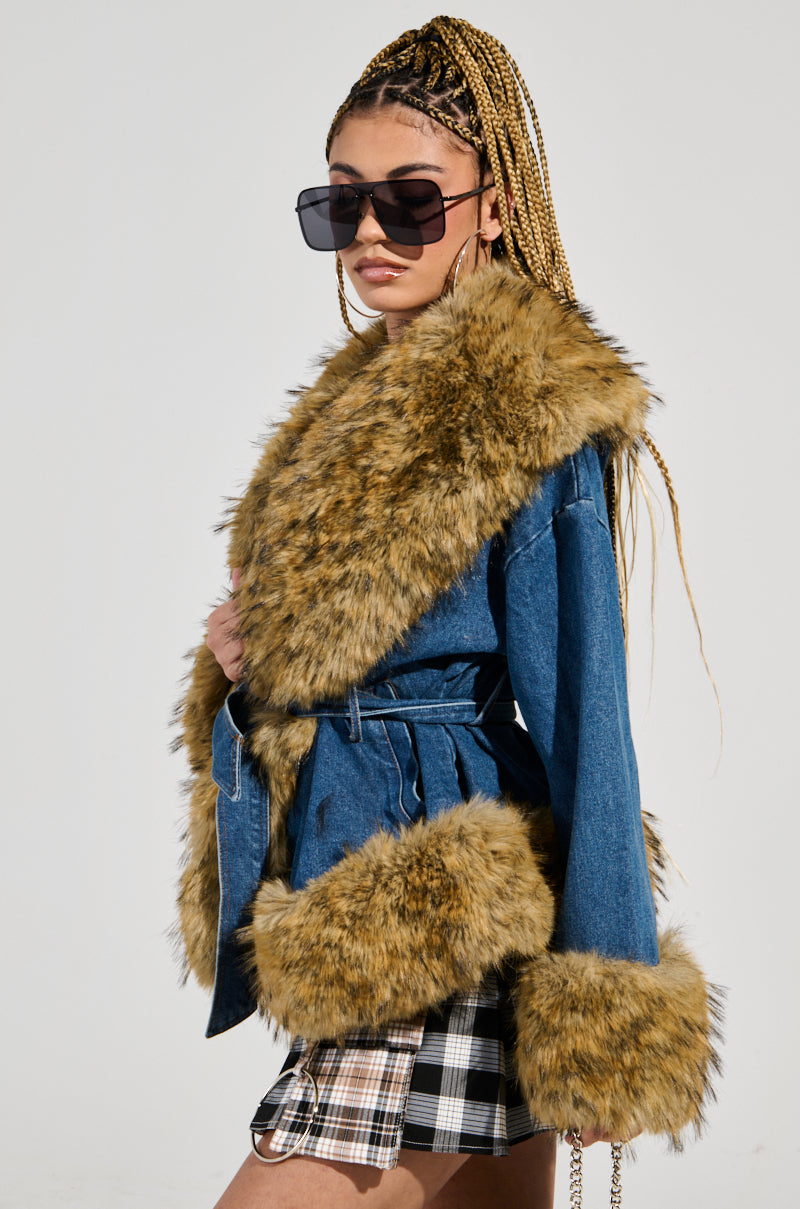 A person confidently poses in oversized sunglasses, sporting the FREDDIE FUR TRIM DENIM MOTO jacket with faux fur trim along the collar and cuffs, and a plaid skirt. Their long braided hair contrasts against a simple, neutral background.