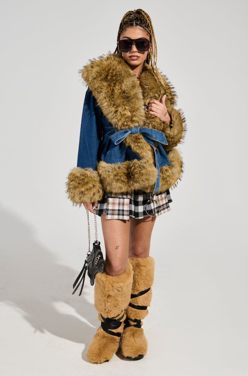 A person models the FREDDIE FUR TRIM DENIM MOTO paired with a plaid skirt and matching fur boots against a plain background. Their braided hair is complemented by sunglasses, and they carry a small black bag featuring an adjustable belt closure.