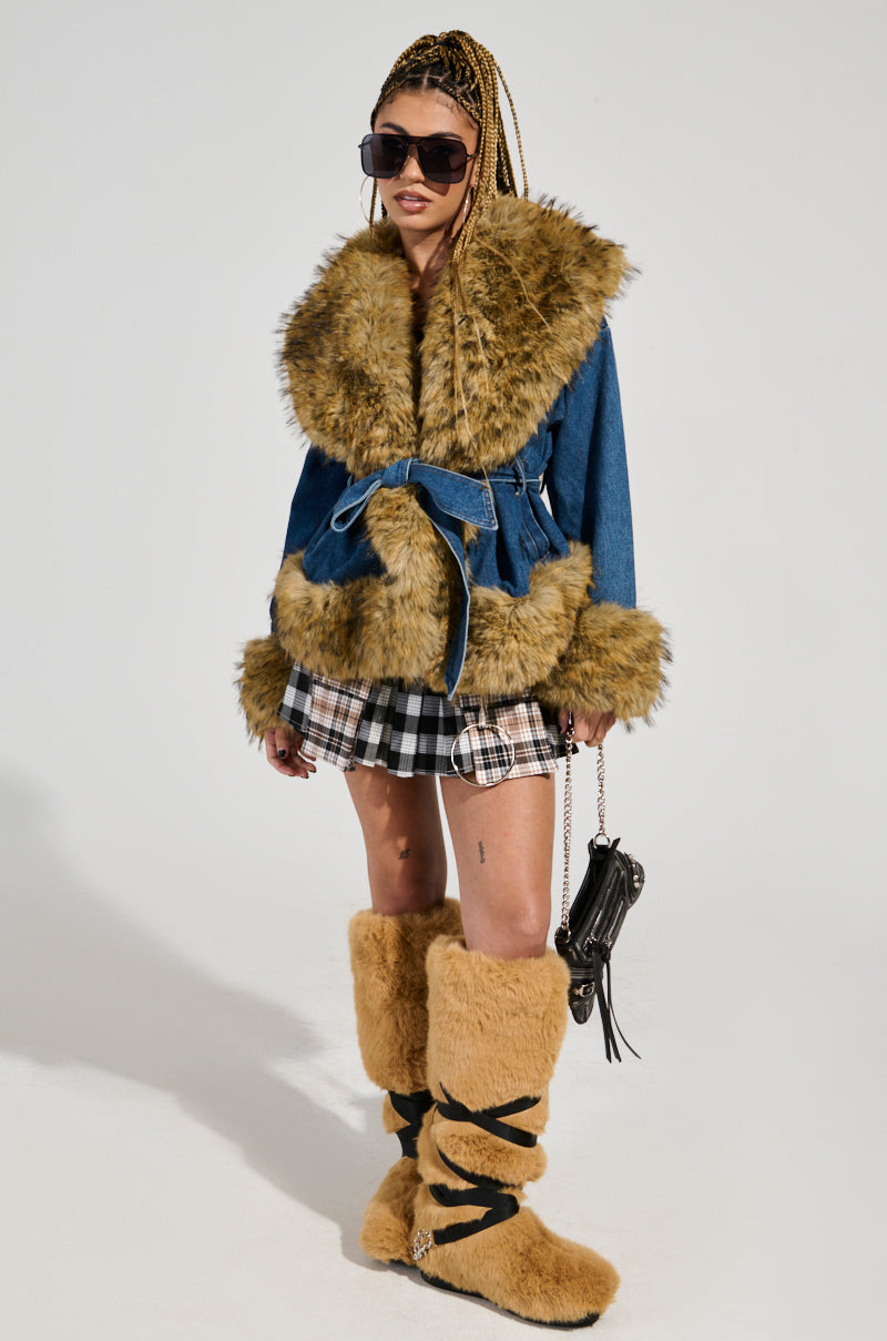 A person dressed in the FREDDIE FUR TRIM DENIM MOTO jacket, paired with a plaid skirt and tall fur boots, carrying a small black handbag. Their long braided hair cascades down as they sport sleek sunglasses.
