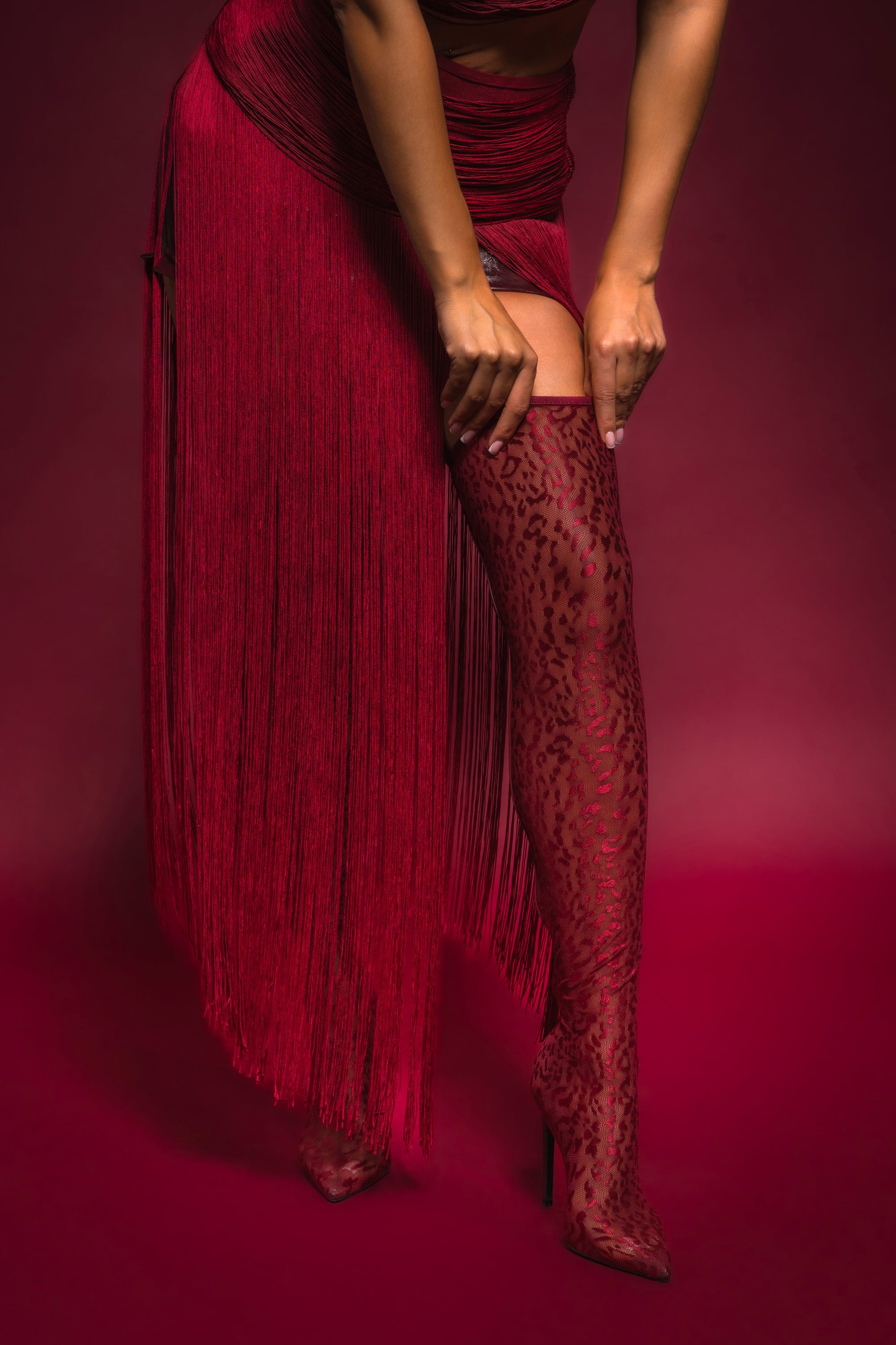 Against a deep red backdrop, someone in a fringed burgundy skirt adjusts the top of an AZALEA WANG NEW FREYJA BURGUNDY LEOPARD LACE STILETTO THIGH HIGH boot; their upper body and face are out of frame. 2