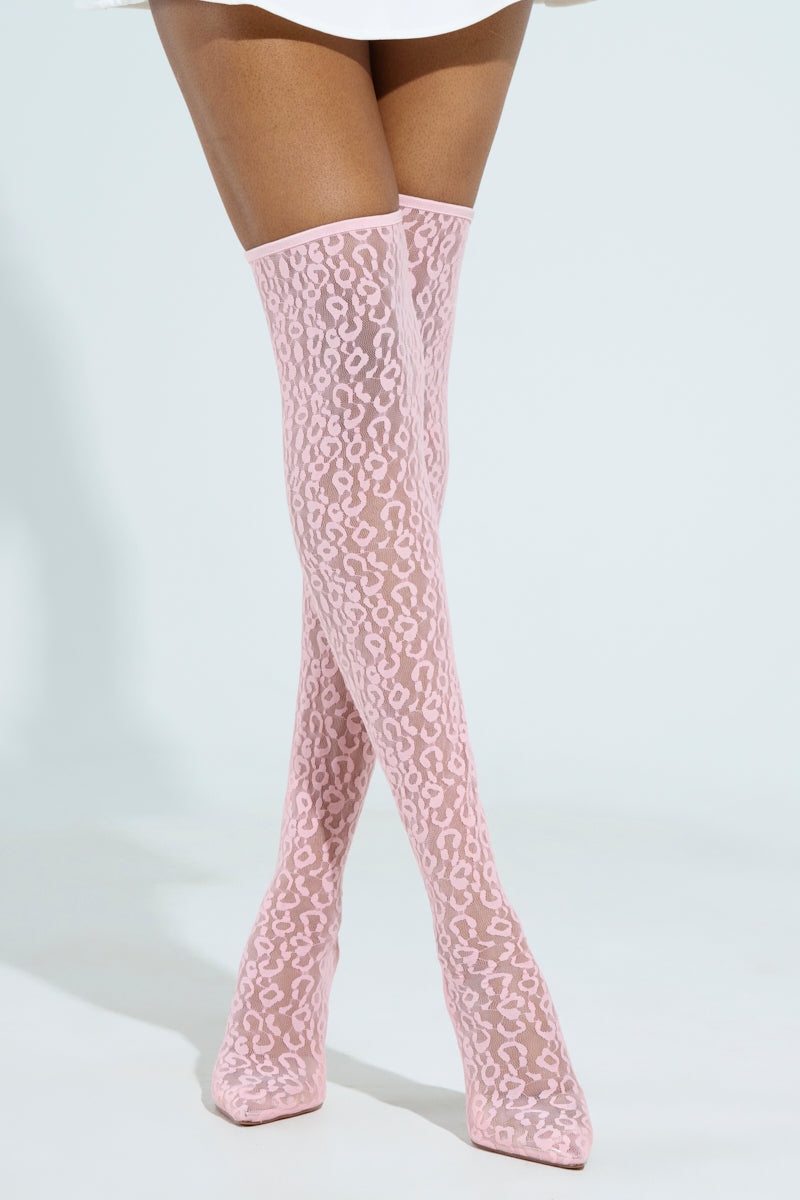 A person crosses their legs while wearing AZALEA WANG FREYJA over the knee lace boots in pink, matched with a white skirt. The background is pale blue.