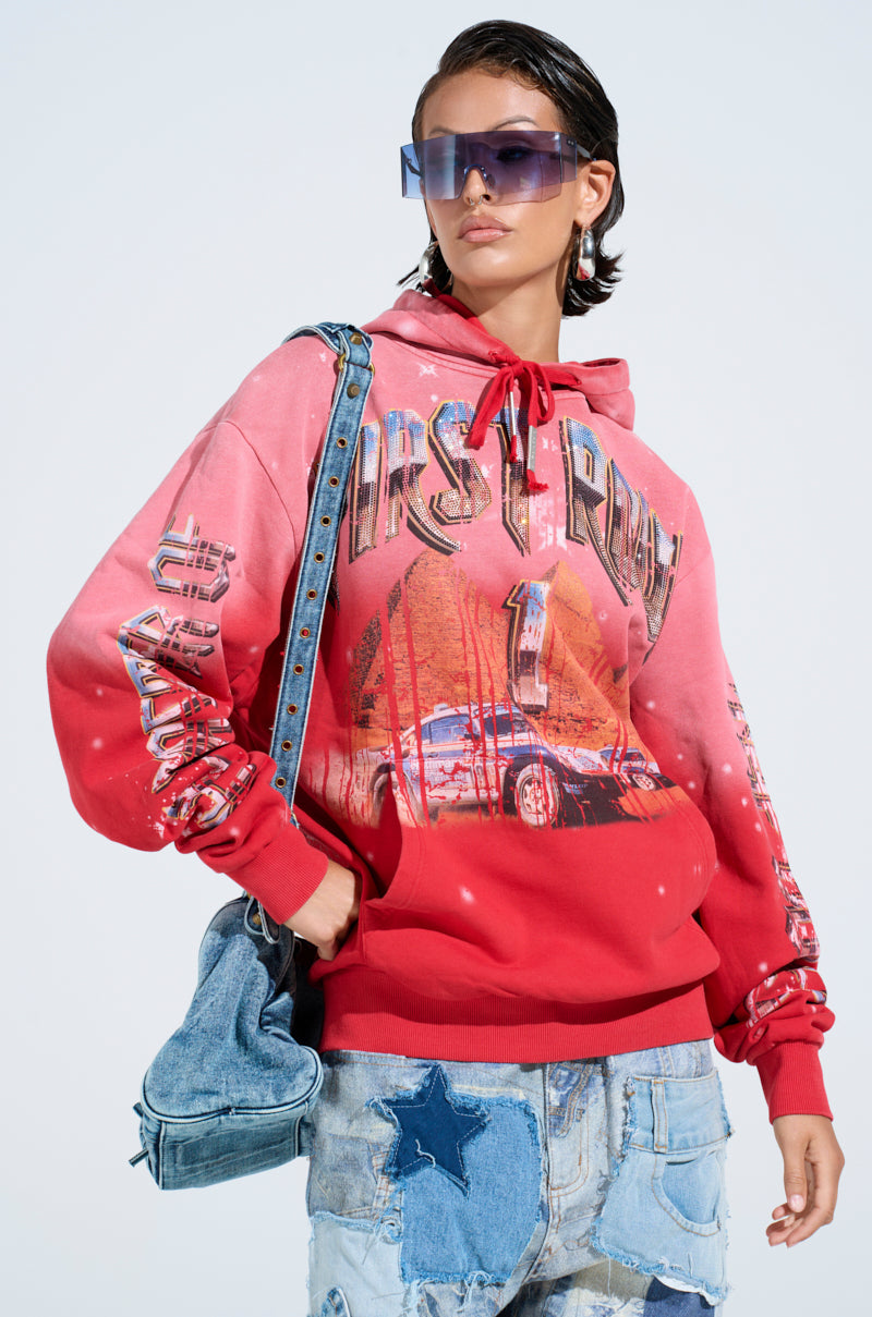 Wearing an ombre red-washed IN THE FAST LANE HOODED SWEATSHIRT with racing graphics, denim shorts with star patches, and oversized blue sunglasses, the individual holds a denim bag against a light background, showcasing their short hair.