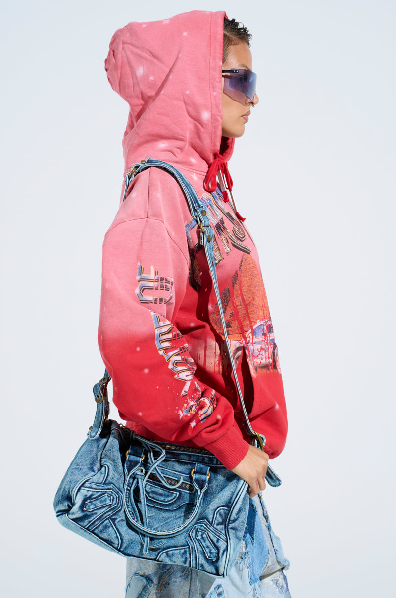A person wearing the "IN THE FAST LANE HOODED SWEATSHIRT" featuring racing-inspired graphics, paired with blue-tinted sunglasses and light blue jeans, carries a textured blue handbag. The backdrop is simple, accentuating their stylish look.