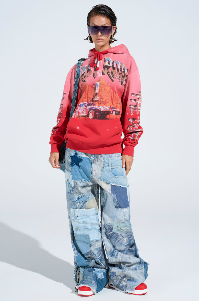 A person wearing the IN THE FAST LANE HOODED SWEATSHIRT in ombre red stands against a plain background with short hair, large sunglasses, oversized patchwork denim jeans, and red and white sneakers.