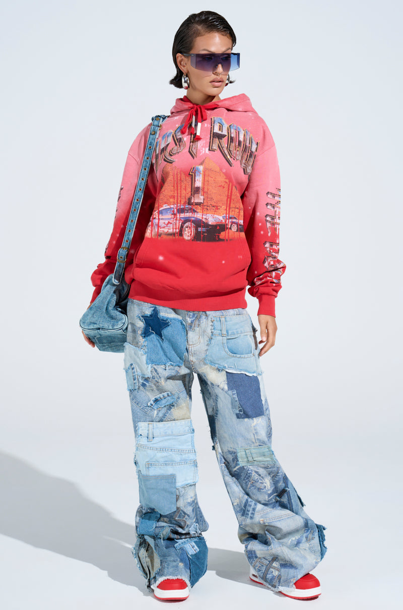 Confidently posing, a person wears the vibrant IN THE FAST LANE HOODED SWEATSHIRT featuring racing graphics. It's paired with patchwork denim jeans, oversized sunglasses, a denim handbag, and red sneakers, all set against a light backdrop.