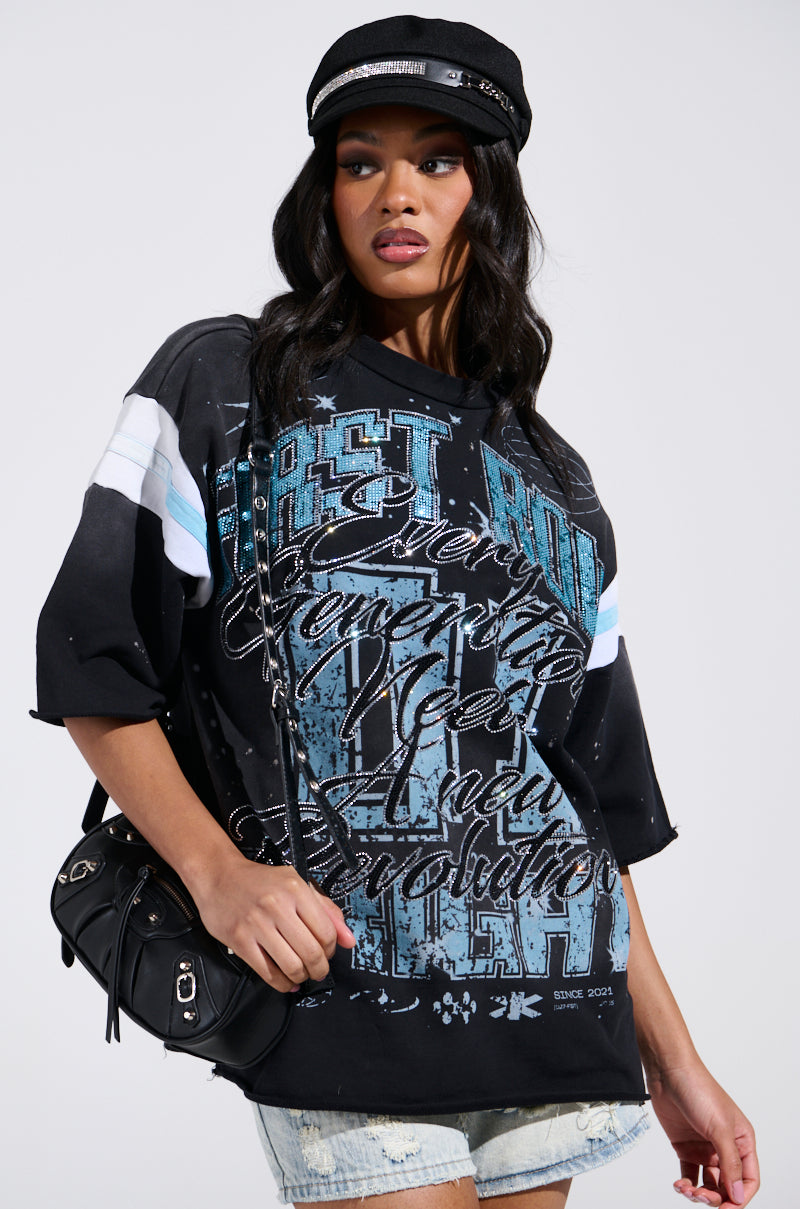 Embodying a grungy-chic style, they wear the BRIGHT FUTURE OVERSIZED GRAPHIC TEE in black with blue accents, holding a small black handbag and sporting distressed denim shorts against a plain backdrop, glancing sideways.